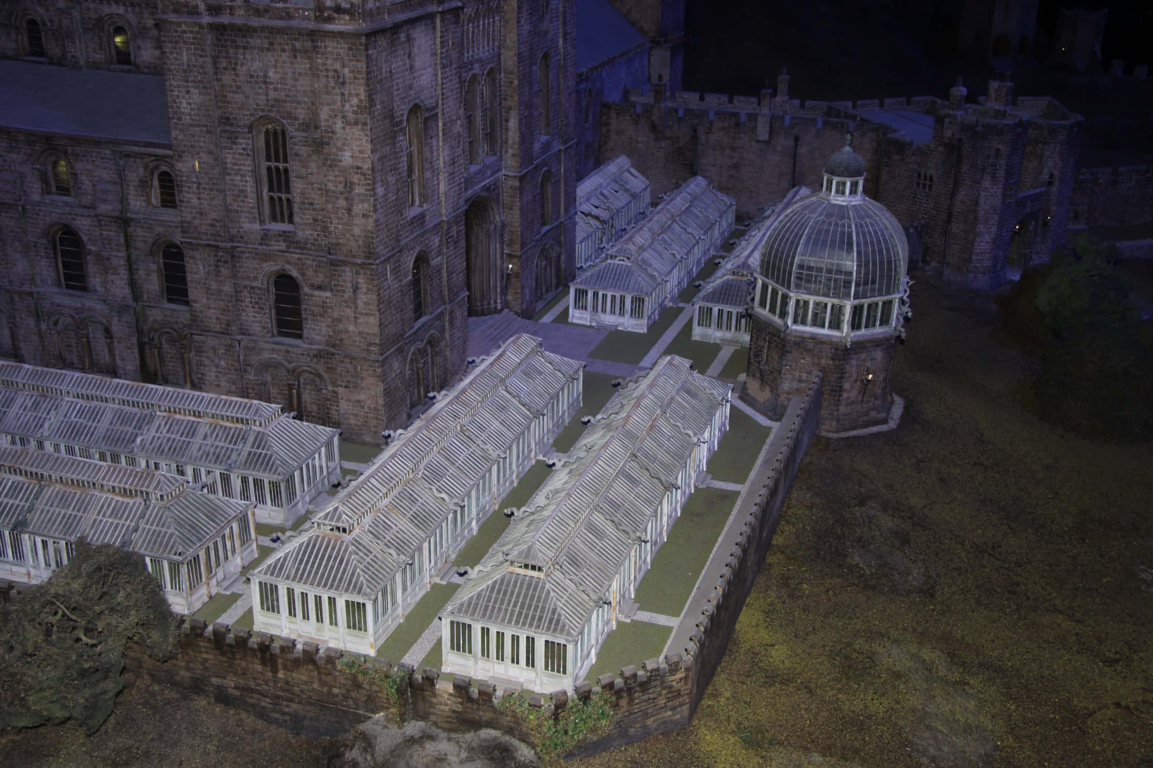 The Hogwarts Greenhouses 桌布 壁紙 — 夜間歷史石城堡建築群的鳥瞰圖，設有照明玻璃溫室和圓頂結構。