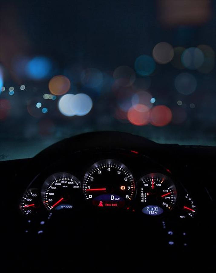 夜間汽車儀表板上的 Speedtest 顯示 壁紙 — 夜間汽車儀表板，紅色指針在散景中模糊的城市燈光映襯下閃爍發光。