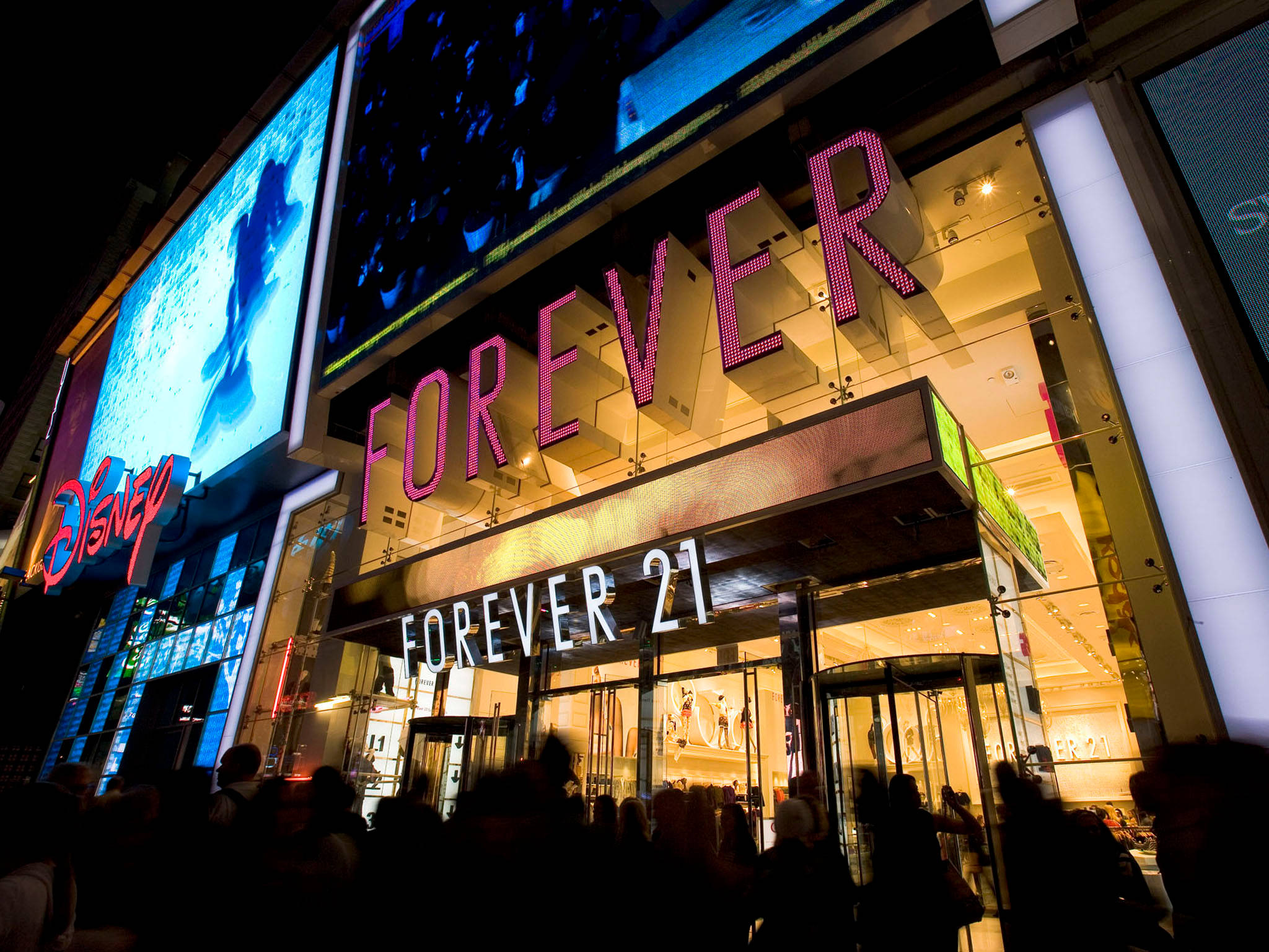 夜間的 Forever 21 服裝店 壁紙 — Forever 21 商店的充滿活力的夜景，帶有發光的粉紅色標牌和上方的迪士尼廣告牌，周圍環繞著人群。