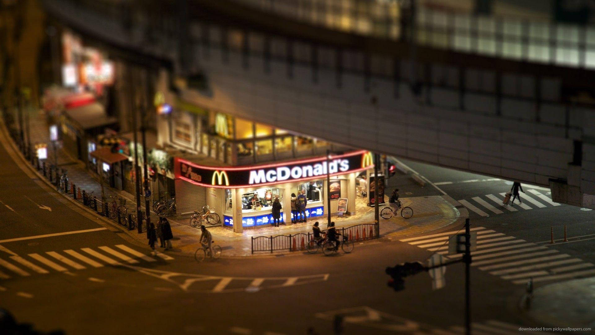 夜間的麥當勞 壁紙 — 天橋下亮燈的麥當勞，彎曲的城市街道上有行人和騎單車人士的微型風格夜景。