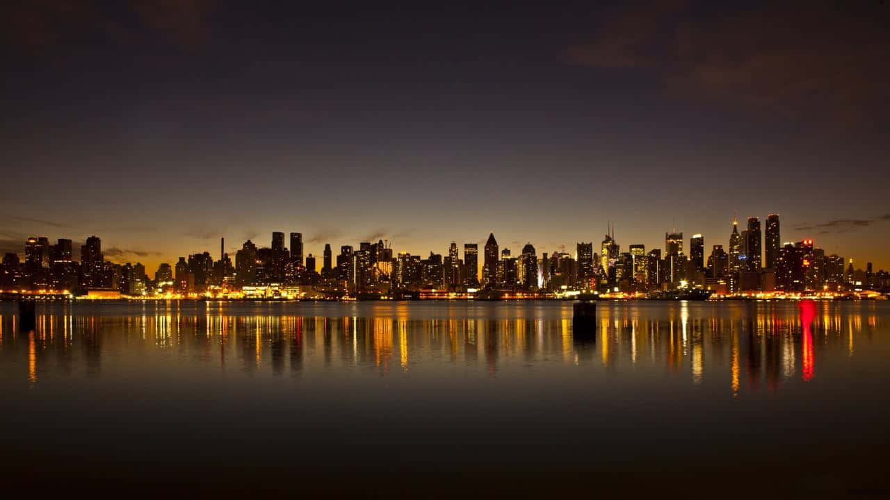 夜間盡享紐約市之美 壁紙 — 令人驚嘆的夜景城市景觀，摩天大樓在暮色天空中閃耀，它們的燈光完美地倒映在平靜的水面上。