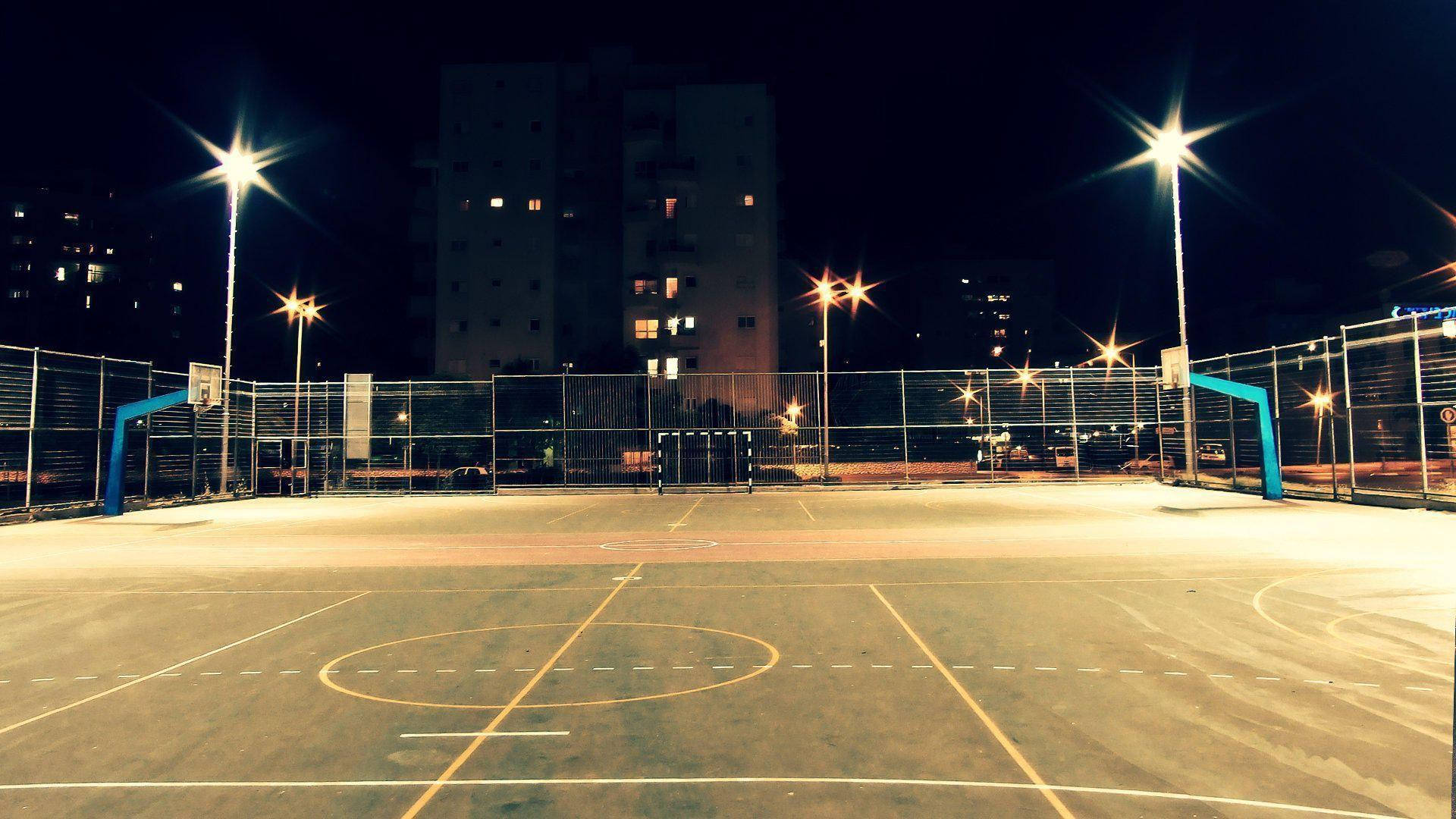 夜間籃球場 壁紙 — 夜晚空無一人的籃球場，被明亮的頭頂燈光照亮，背景是公寓大樓。