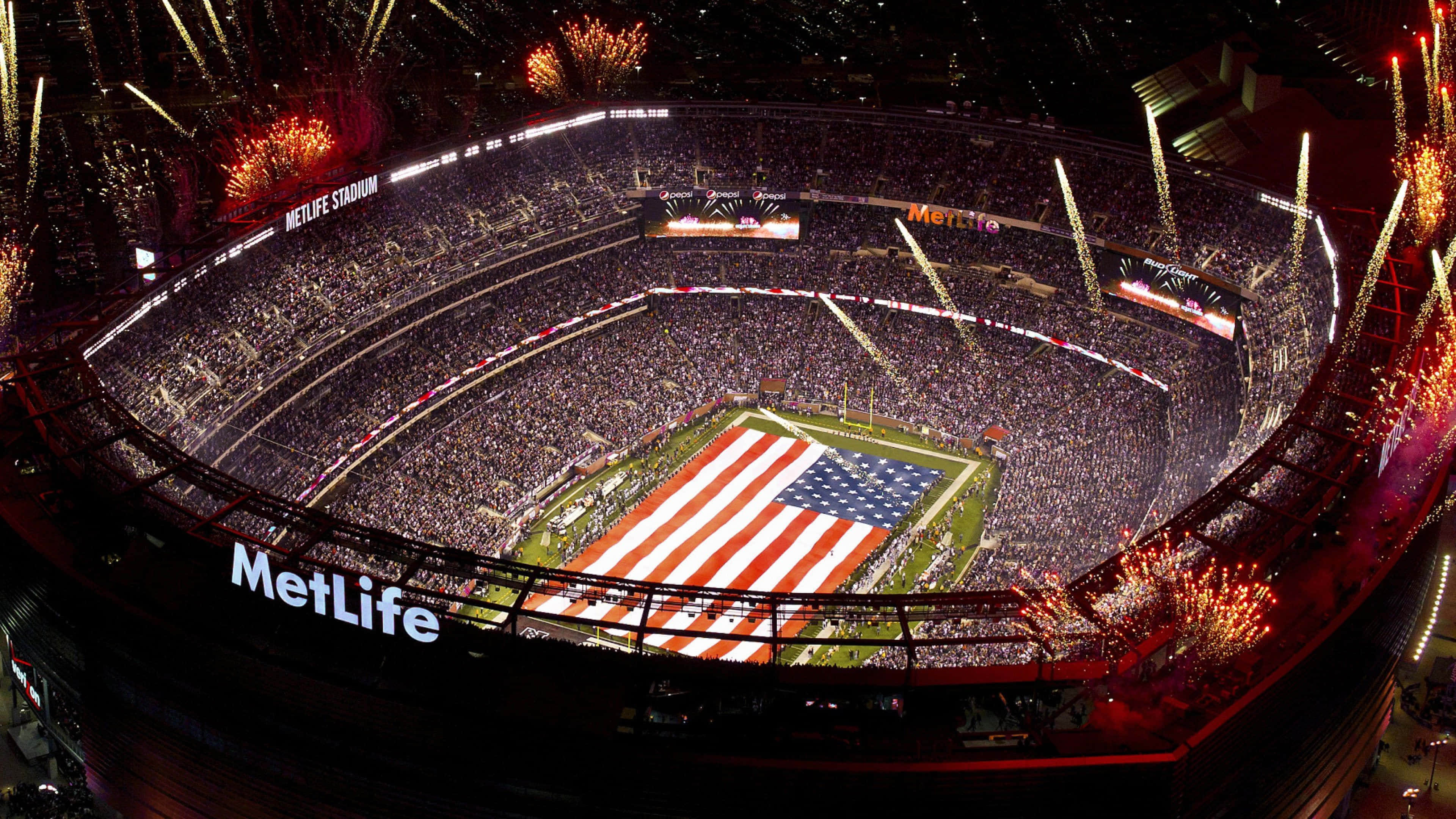 Super Bowl 桌布 壁紙 — 夜間美式足球場MetLife Stadium的鳥瞰圖，場內擠滿球迷，球場上空煙花綻放，顯示一面巨大的美國國旗。