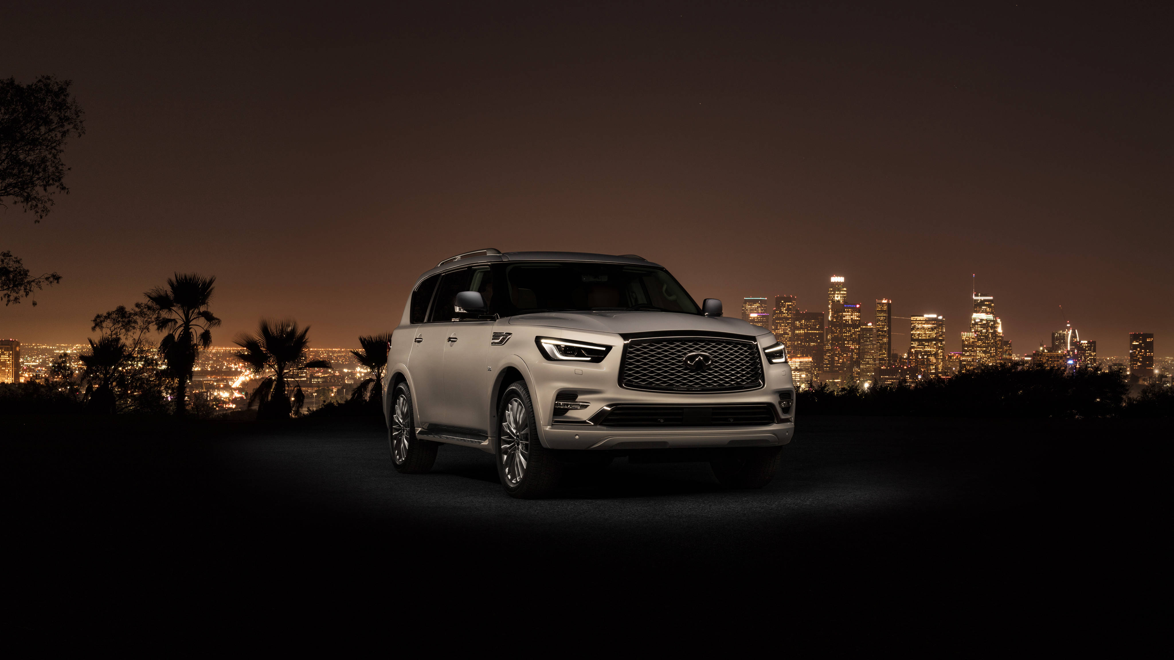 夜間英菲尼迪 QX80 壁紙 — 一輛銀色 Infiniti QX80 SUV 停泊在山丘上，俯瞰著夜晚充滿活力的城市天際線，前景有棕櫚樹的剪影。