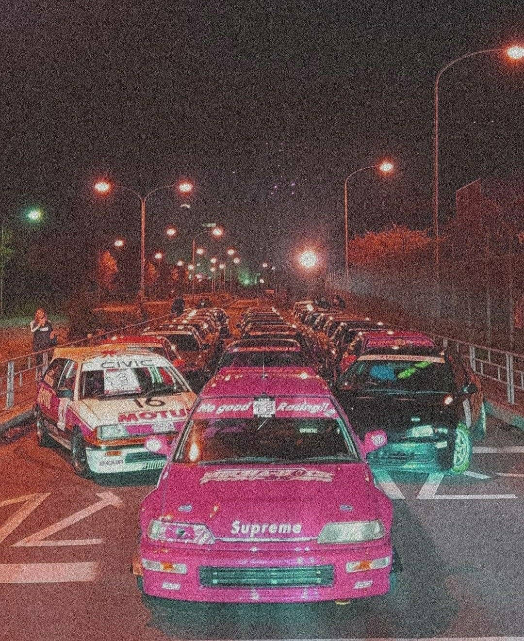 帶有 Supreme 標誌的粉紅色賽車在夜晚與其他改裝車一起停放。