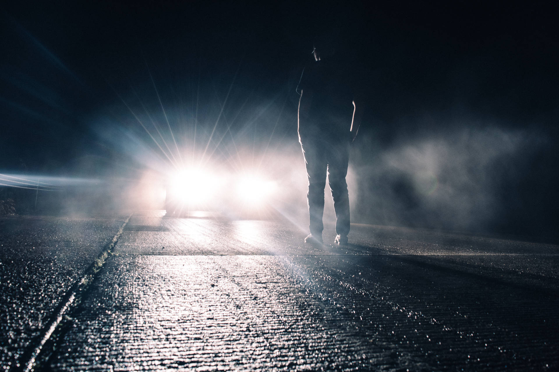 一個人的剪影站在夜晚潮濕的道路上，車頭燈閃爍。