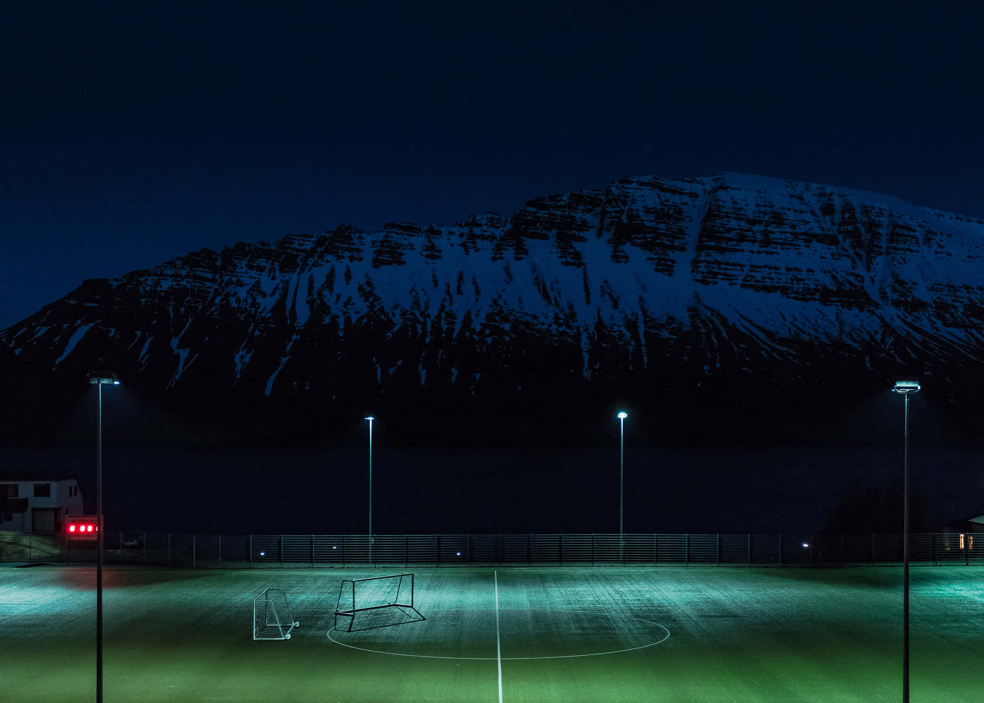 夜晚燈光照亮的空蕩足球場，背景是積雪的山峰。