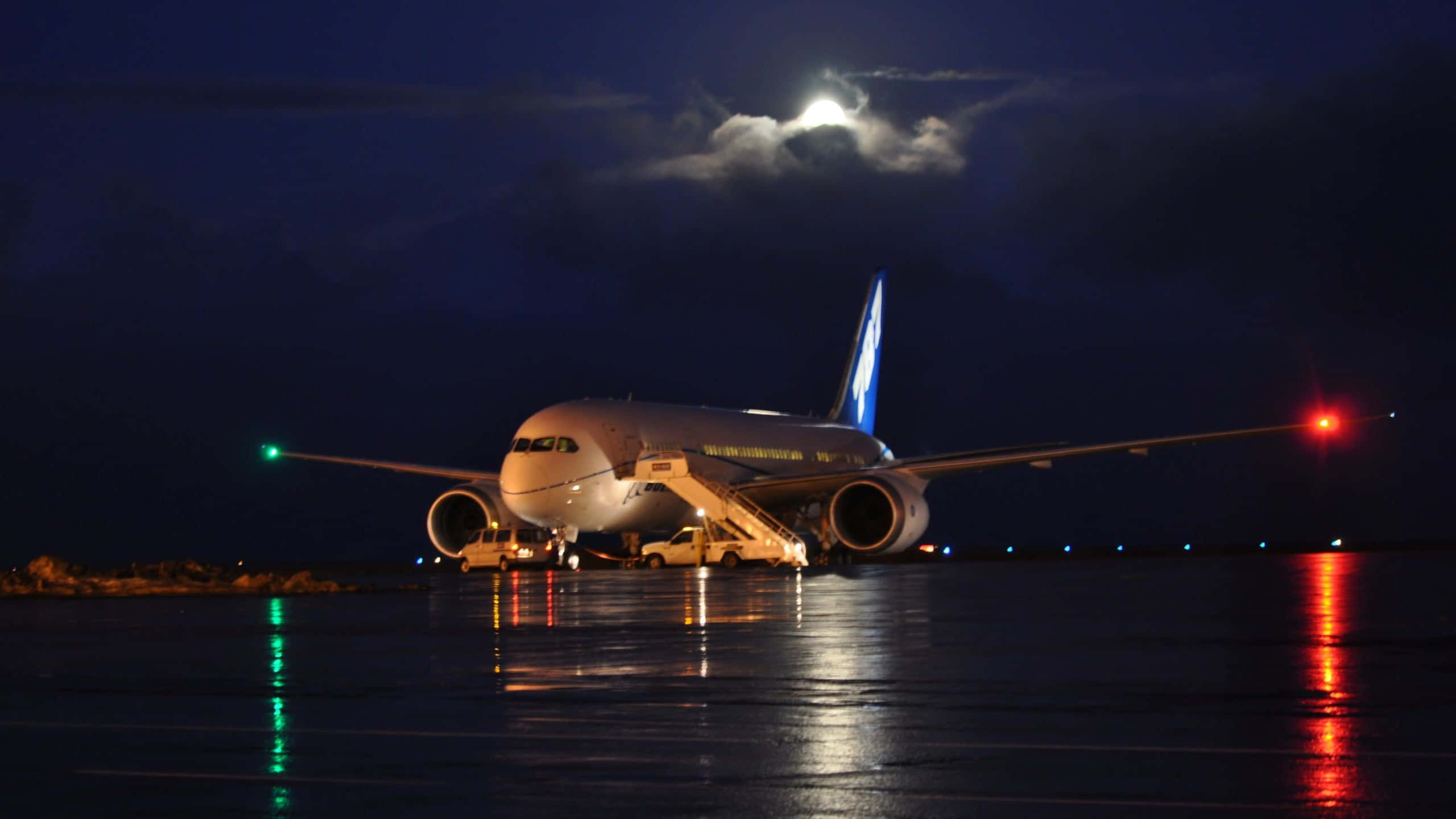 夜間跑道上的飛機與航行燈 1440p 小型飛機背景 壁紙 — 一架大型飛機在夜間停泊在濕滑的停機坪上，明亮的月亮和雲朵下，紅色和綠色的燈光反射在地面上。