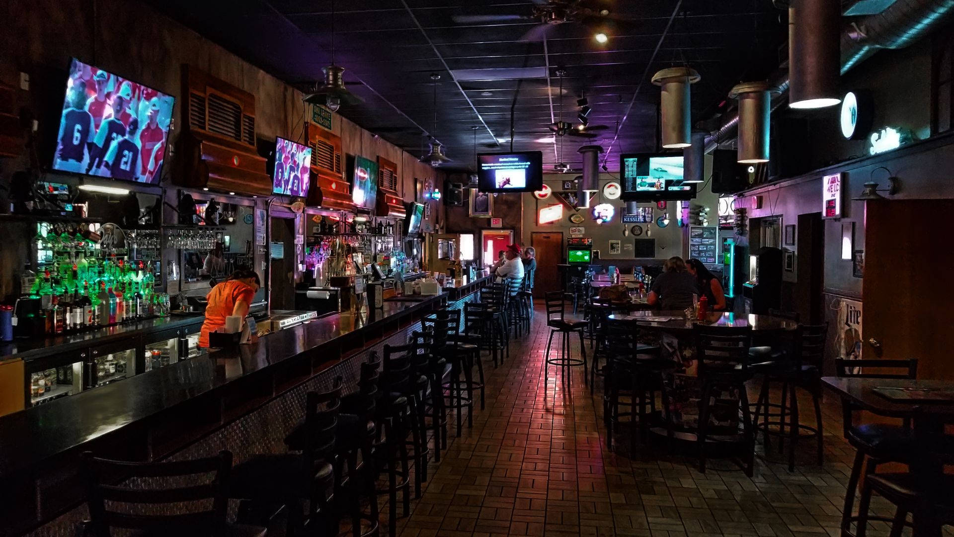 夜間酒館攝影 壁紙 — 燈光昏暗的運動酒吧，長吧檯上方有發光的電視屏幕，顧客坐在霓虹燈下的桌子旁。