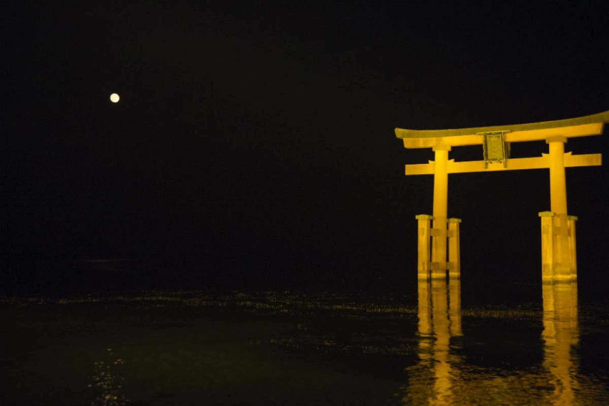 夜間鳥居門 壁紙 — 一座發光的黃色鳥居矗立在黑暗的水中，明亮的月亮投下寧靜的倒影。