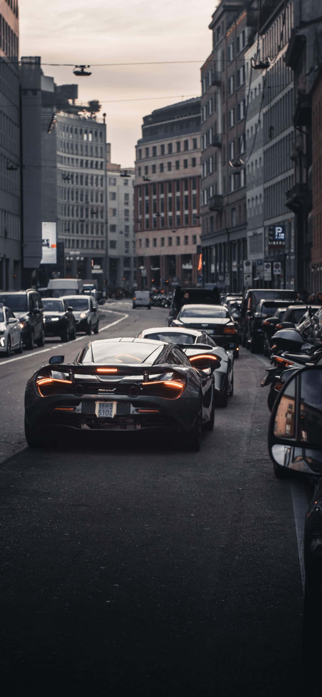 夢想中的奢華跑車：iPhone Xs Max McLaren 720s 壁紙 — 一輛時尚的黑色跑車帶有發光的橙色尾燈，在狹窄的城市街道上行駛，兩旁是高樓大廈和停放的車輛，天空陰沉。