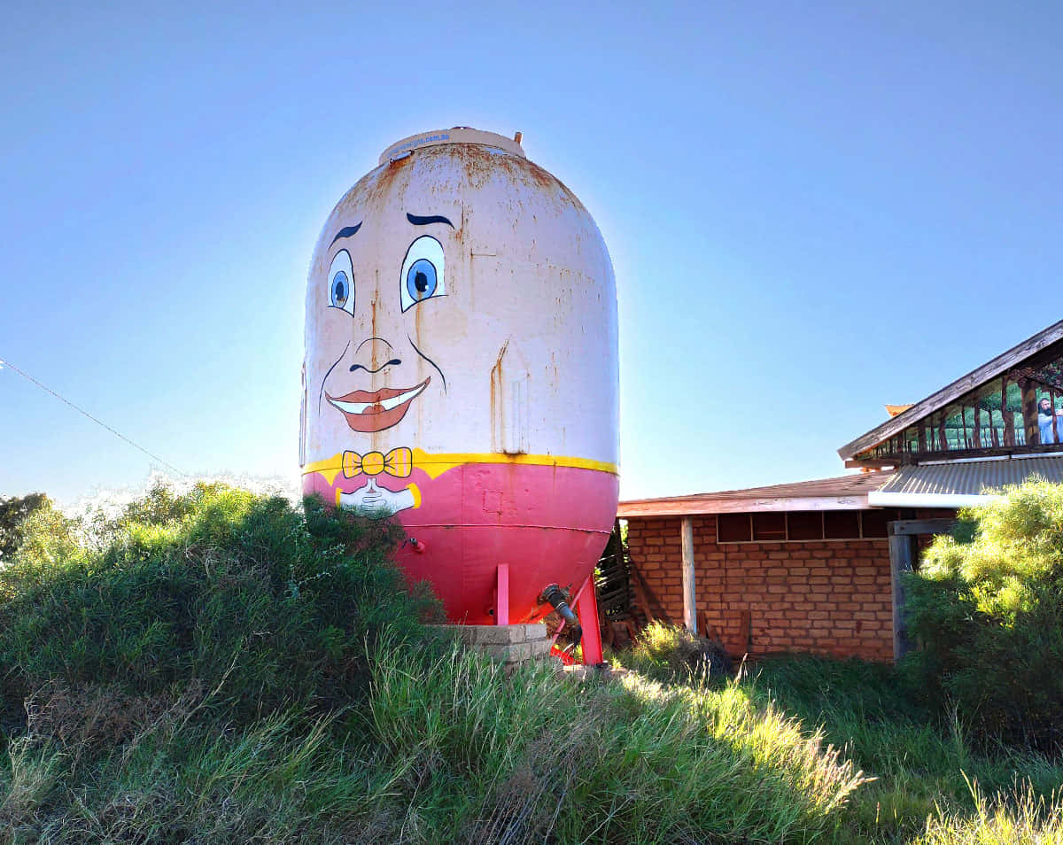 大型 Humpty Dumpty 角色圖像 壁紙 — 一個巨大的生鏽筒倉，上面畫著一張開心的卡通臉，矗立在藍天白雲下的高草叢中。