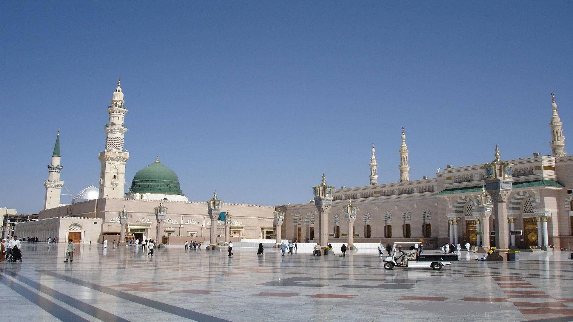 Madina Sharif 桌布 壁紙 — 大型清真寺，綠色圓頂和宣禮塔，在晴朗藍天下，人們在反射廣場上行走。