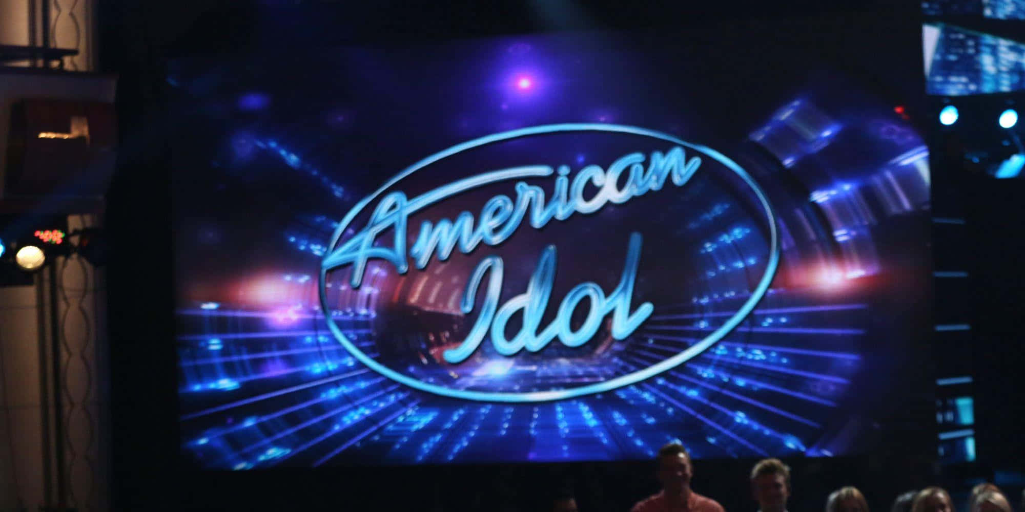 american idol 桌布 壁紙 — 大屏幕顯示發光的American Idol標誌，藍色和紫色的舞台燈光向外輻射。