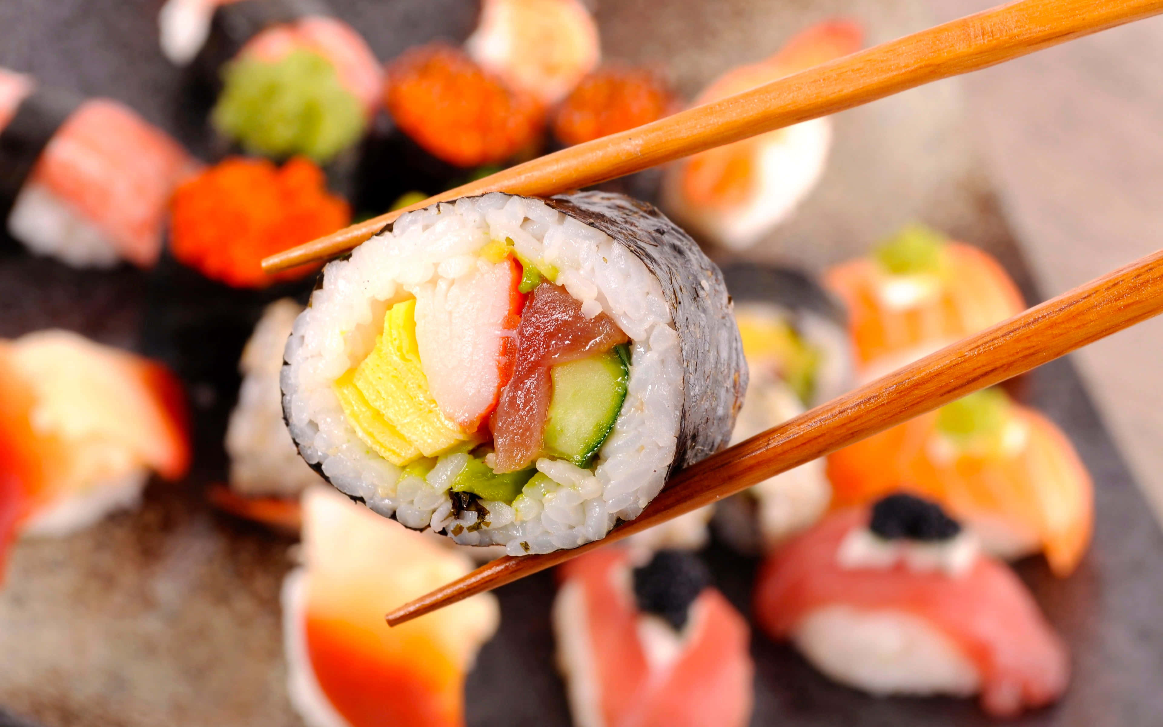 大捲壽司圖片 壁紙 — 筷子夾住壽司卷（內含米飯、吞拿魚、青瓜和雞蛋）的特寫，背景是模糊的各式壽司。