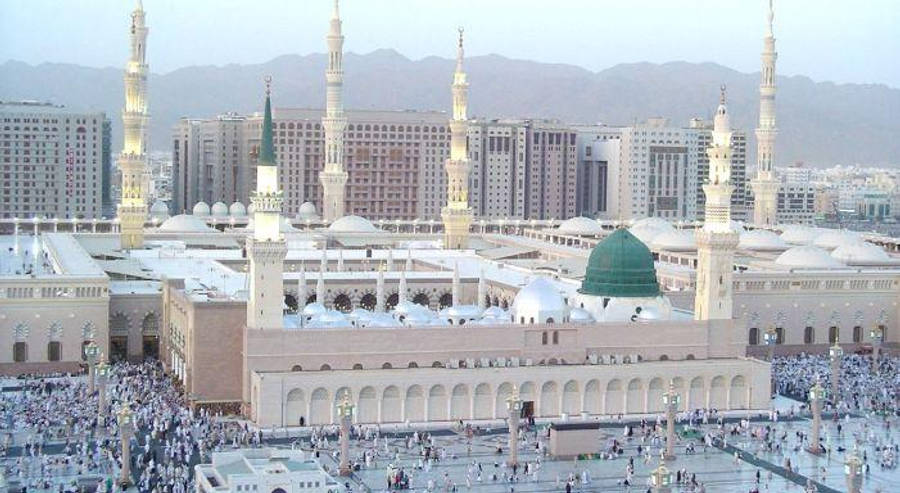 Madina 桌布 壁紙 — 大清真寺的鳥瞰圖，擁有綠色圓頂和高聳的宣禮塔，周圍環繞著人群和現代建築，背景是朦朧的山脈。