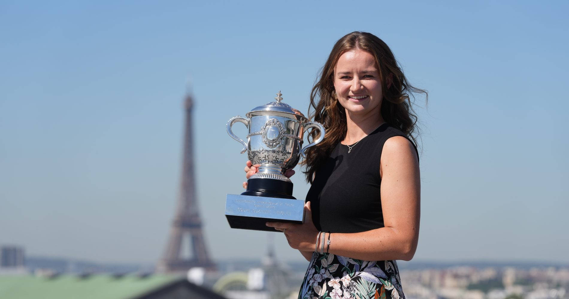 大滿貫單打冠軍巴博拉·克雷奇科娃 壁紙 — 一位微笑的黑上衣女士手持銀色獎盃，背景是晴朗的藍天和艾菲爾鐵塔。