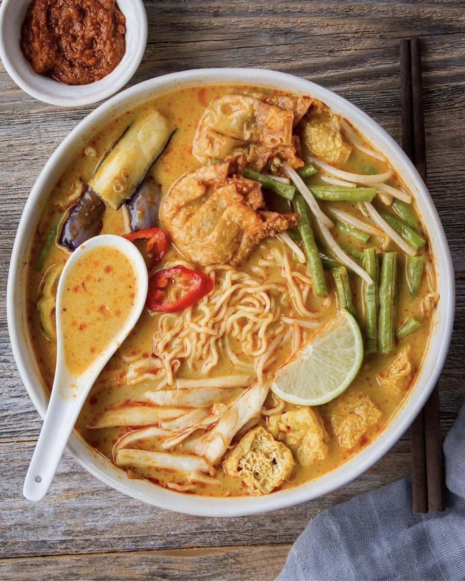 大碗咖喱叻沙 壁紙 — 一碗熱騰騰的濃郁黃咖哩湯麵，配上豆腐、豆芽、青檸和辣椒，放在質樸的木桌上。