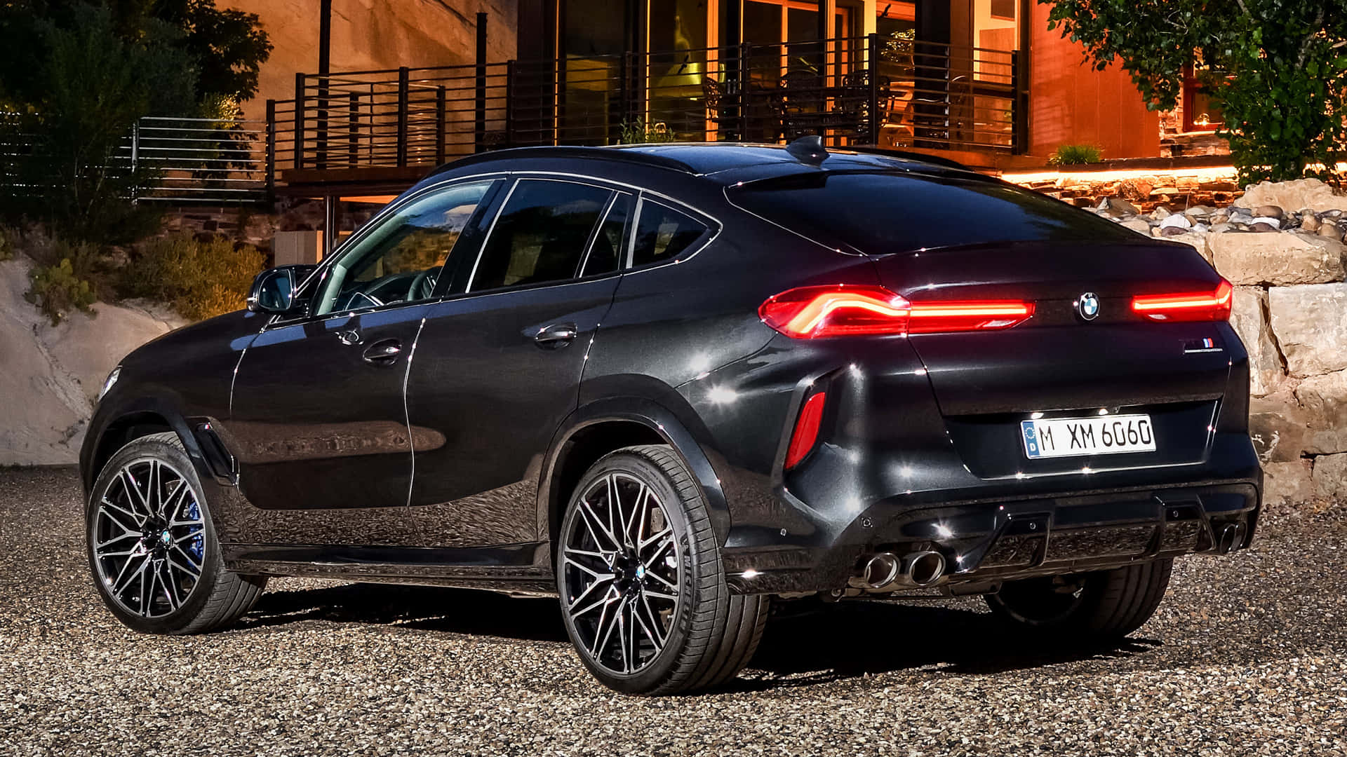 大膽奢華的BMW X6 M 壁紙 — 一輛時尚的黑色BMW X6M停在黃昏的碎石路上，尾燈閃爍，背景是一座現代住宅。