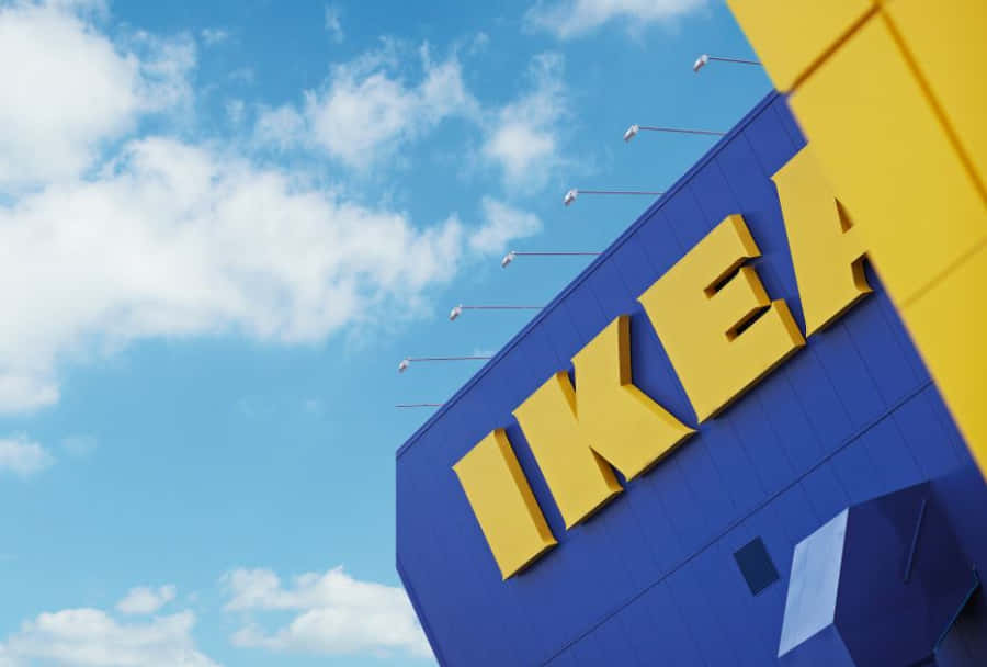 Ikea 桌布 壁紙 — 大膽的黃色IKEA字母在藍色建築上閃耀，背景是明亮的藍天和蓬鬆的白雲。