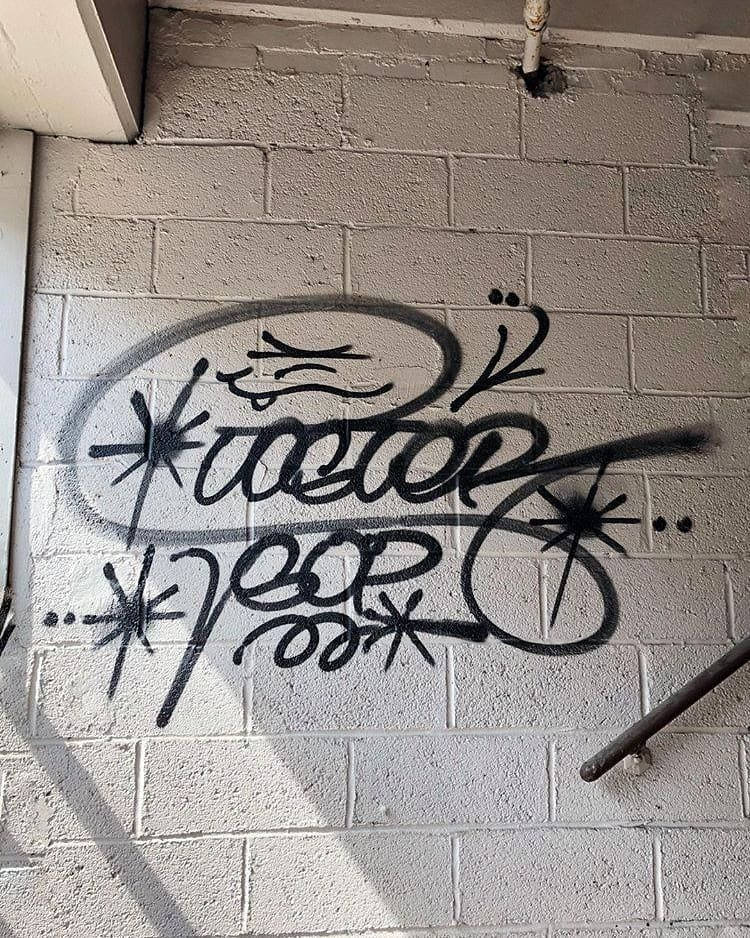 Black And White Graffiti 桌布 壁紙 — 大膽的黑色塗鴉標籤，帶有星爆圖案，噴塗在簷篷下有紋理的白色磚牆上。