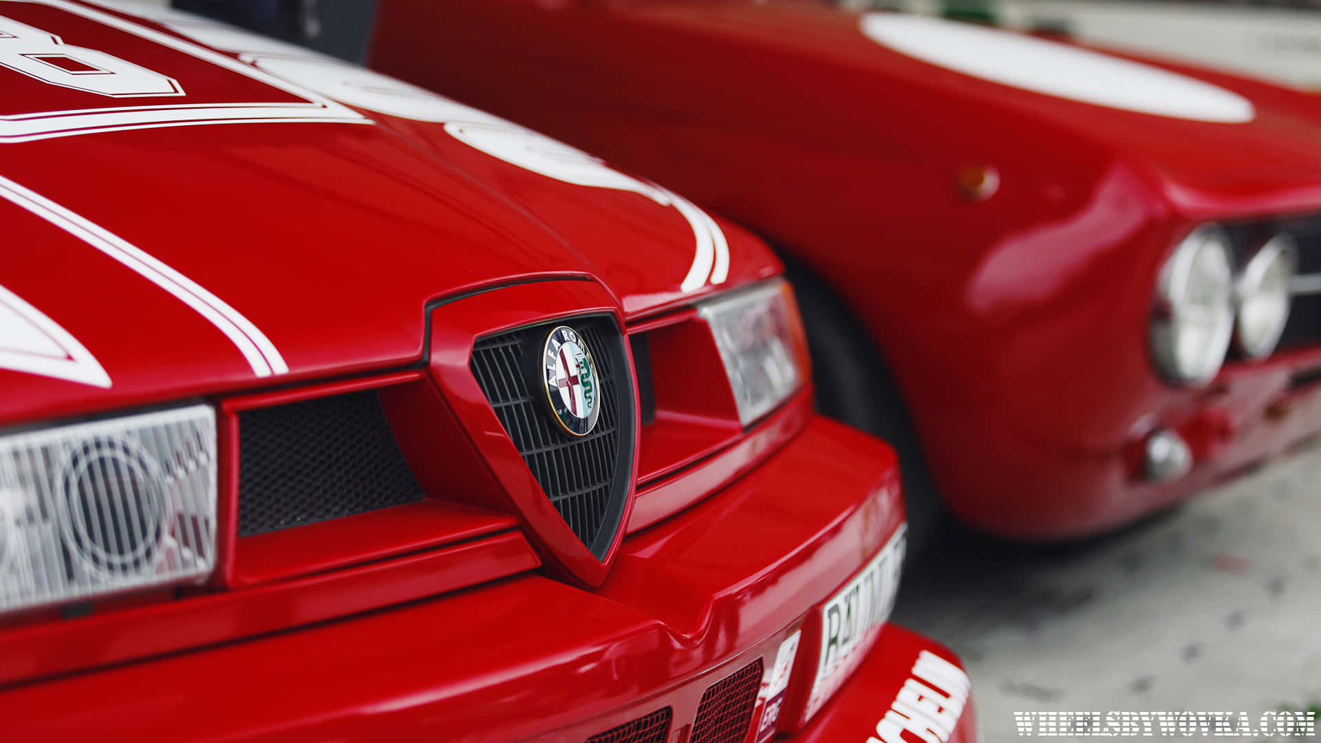 大膽而傳奇的Alfa Romeo 壁紙 — 車庫中兩輛帶有白色條紋和標誌性盾牌的亮紅色愛快羅密歐賽車的特寫。