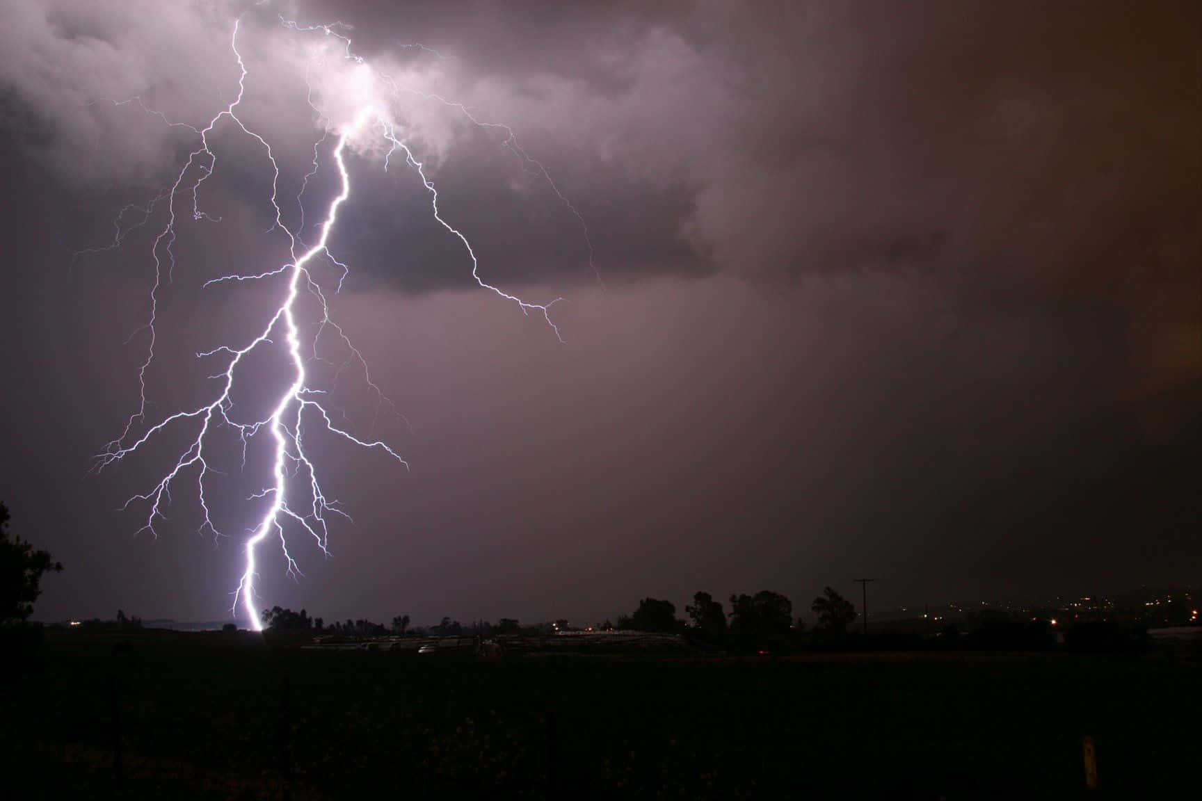 大自然怒火的力量 壁紙 — 一道巨大的閃電在暴風雨的夜空中分叉，下方是黑暗的鄉村景觀，遠處有城市燈光。
