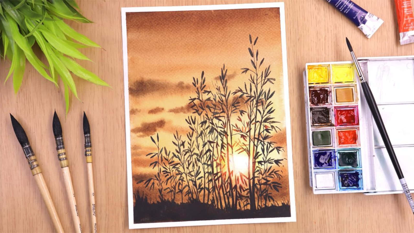 大自然的藝術 壁紙 — 一幅水彩畫，描繪竹子在溫暖日落天空下的剪影，周圍是木桌上擺放的藝術用品。