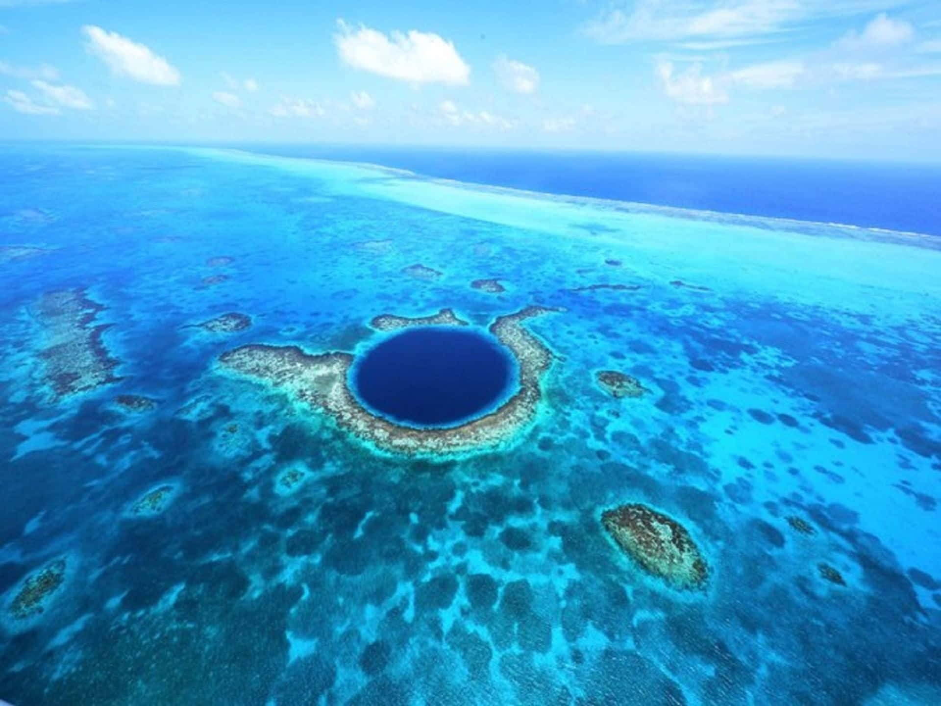 大藍洞與廣闊海洋的海景 壁紙 — 一個壯觀的圓形藍洞的鳥瞰圖，周圍環繞著充滿活力的綠松石珊瑚礁，在明亮的藍天下有零星雲朵。