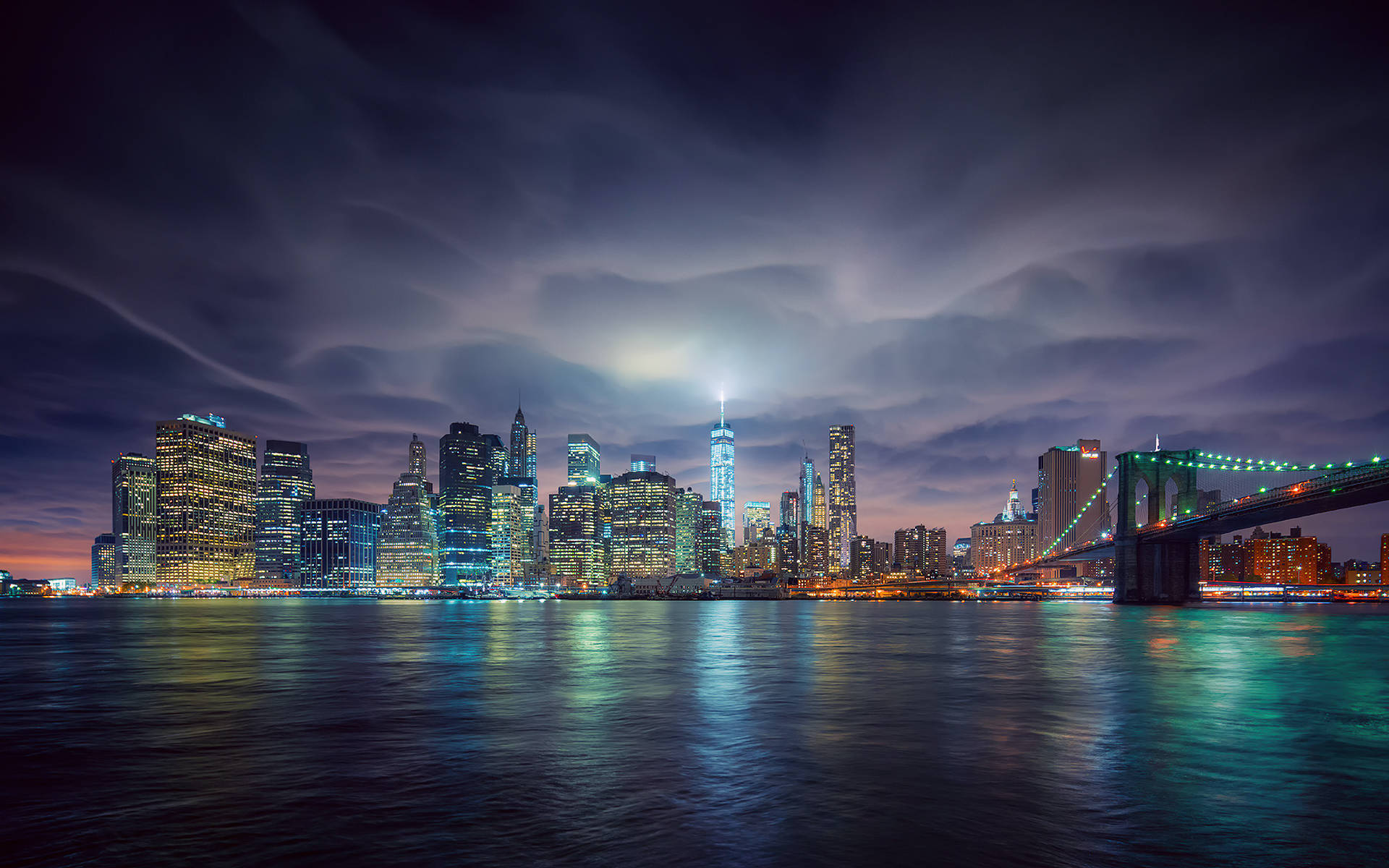 大蘋果城市燈光 壁紙 — 紐約市天際線的壯麗夜景，照亮摩天大樓和布魯克林大橋在平靜的水面上倒映，天空有戲劇性的雲層。