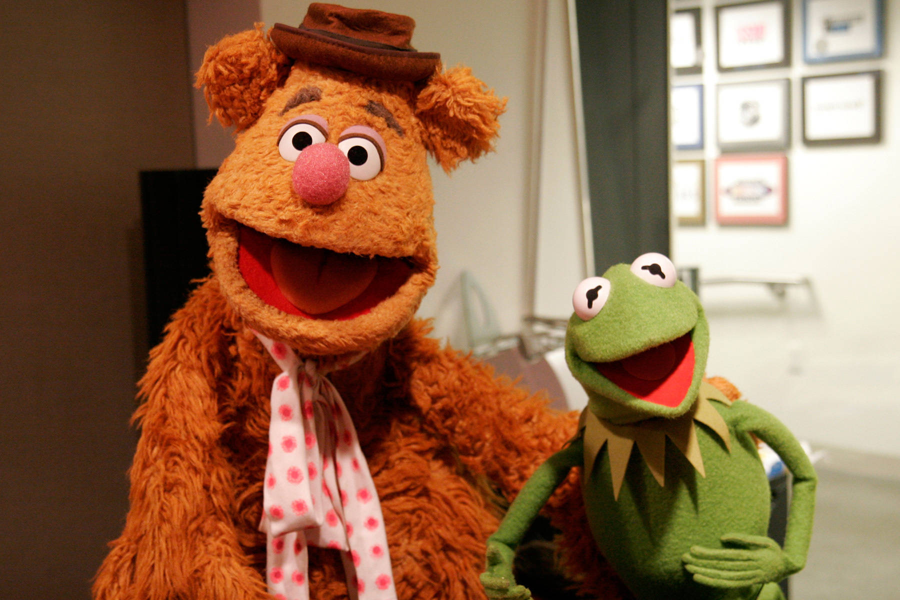 大號Fozzie Bear與Kermit Frog 壁紙 — Fozzie Bear 戴著棕色帽子和圓點圍巾，在溫暖燈光的室內環境中，與Kermit the Frog一起微笑。