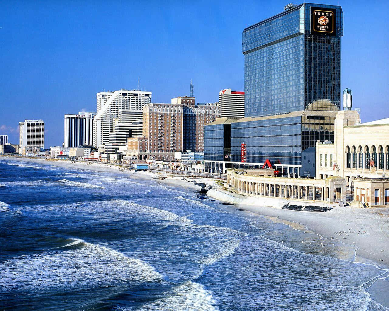 大西洋城絢麗日落 壁紙 — 充滿活力的沿海城市景觀，高聳的酒店和賭場沿著沙灘排列，海浪在清澈的藍天下拍打。