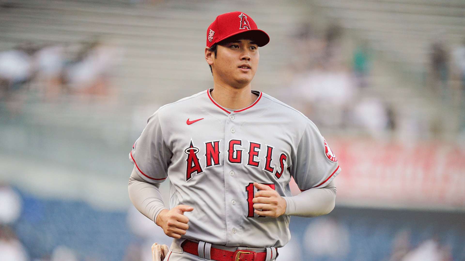 大谷翔平場上慢跑 壁紙 — 身穿天使隊球衣的棒球選手在球場上慢跑，背後是模糊的體育館人群。