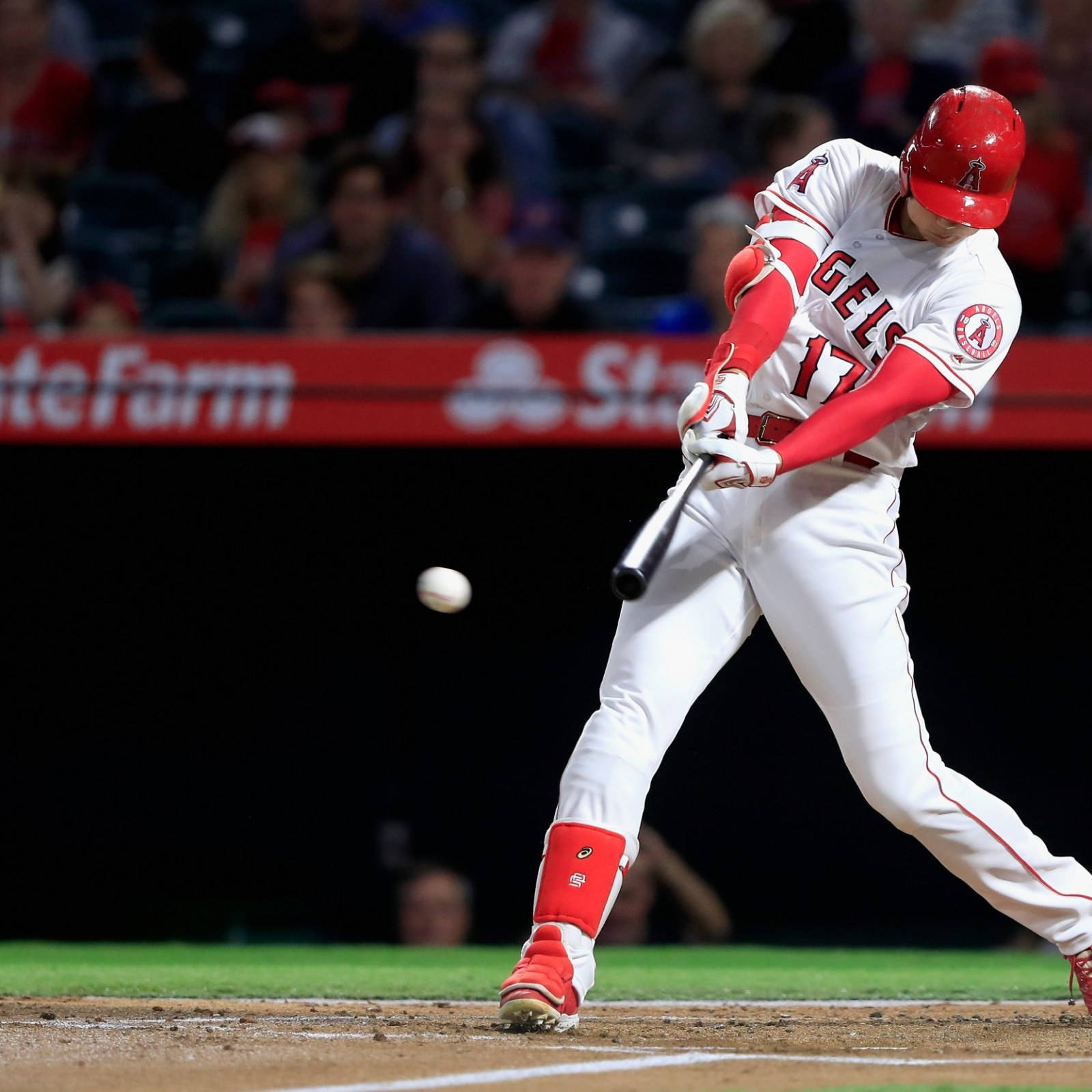大谷翔平揮棒擊球瞬間 壁紙 — 一名身穿紅白球衣的棒球手在夜間比賽中揮棒擊球，背景是模糊的人群。