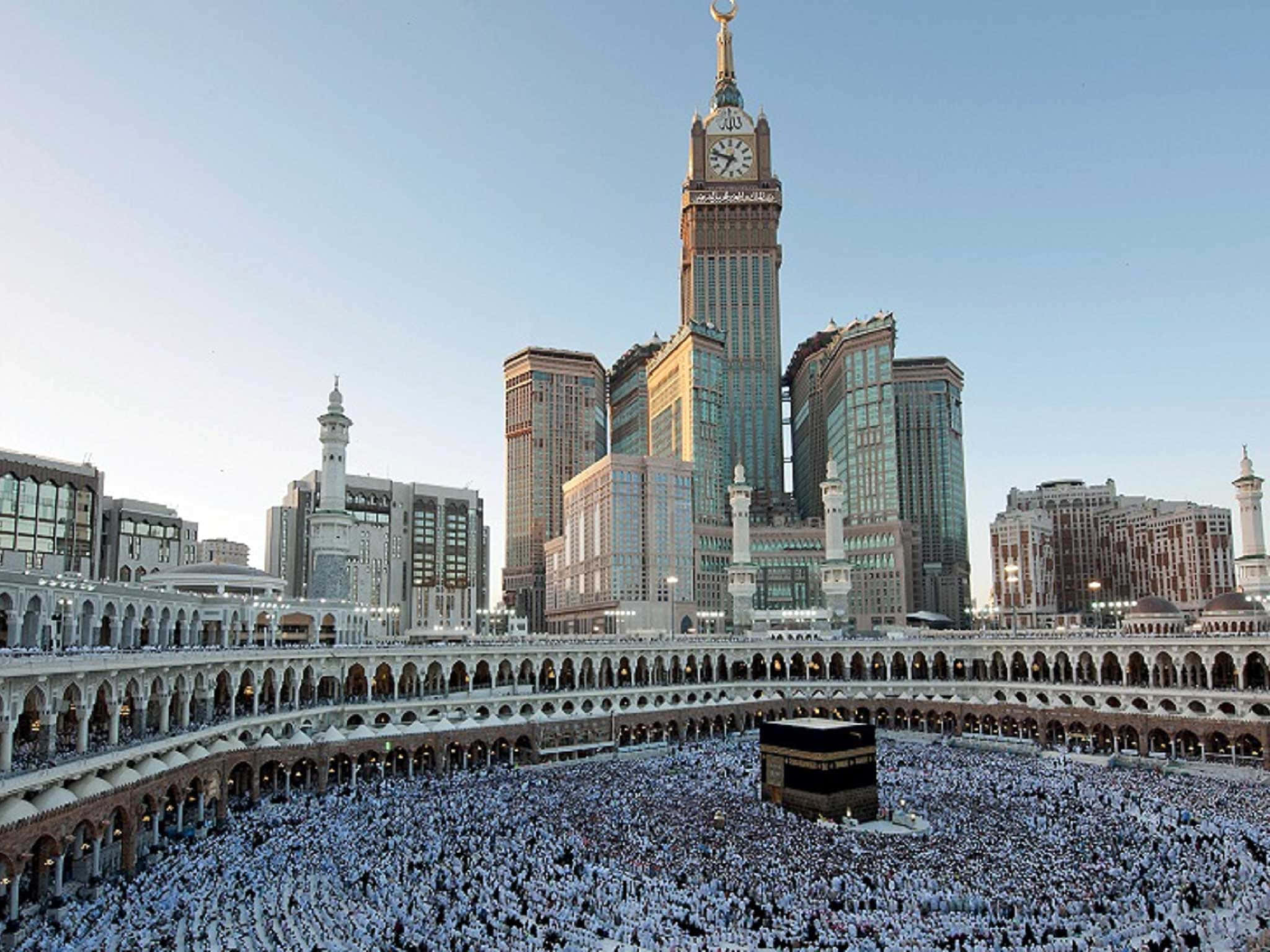 Makkah 桌布 壁紙 — 大量身穿白衣的人群在晴朗的天空下圍繞著卡巴天房，背景是現代摩天大樓和一座鐘樓。