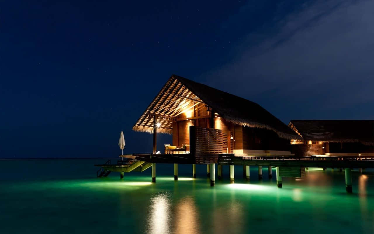 大開曼島的壯麗海灘 壁紙 — 夜晚中發光的水上平房，溫暖的燈光在星空下反射在碧綠色的水面上。