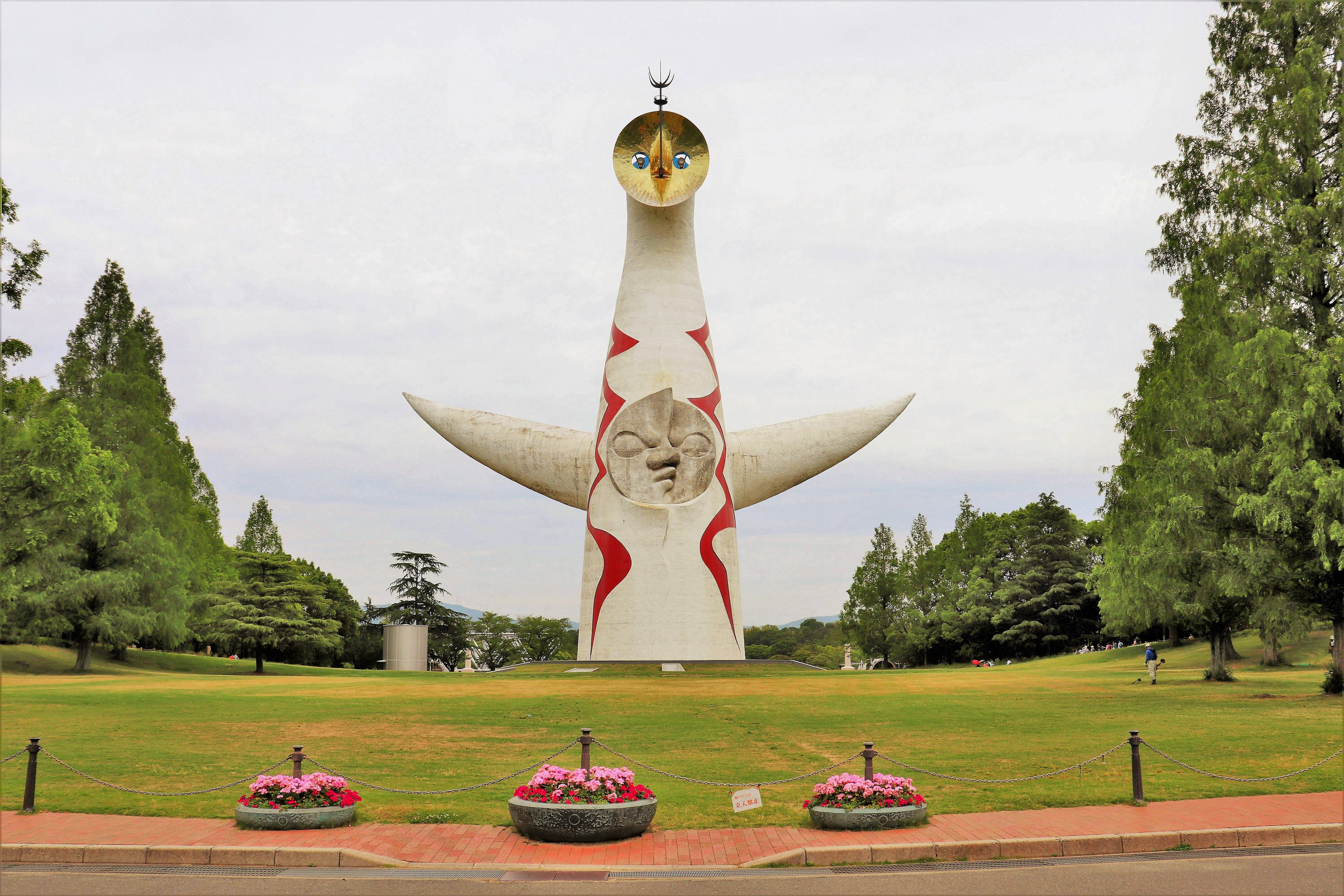 大阪世博'70公園 壁紙 — 一座高聳的白色雕塑，帶有紅色點綴和風格化的面孔，矗立在陰天下一片鬱鬱蔥蔥的綠色公園中。