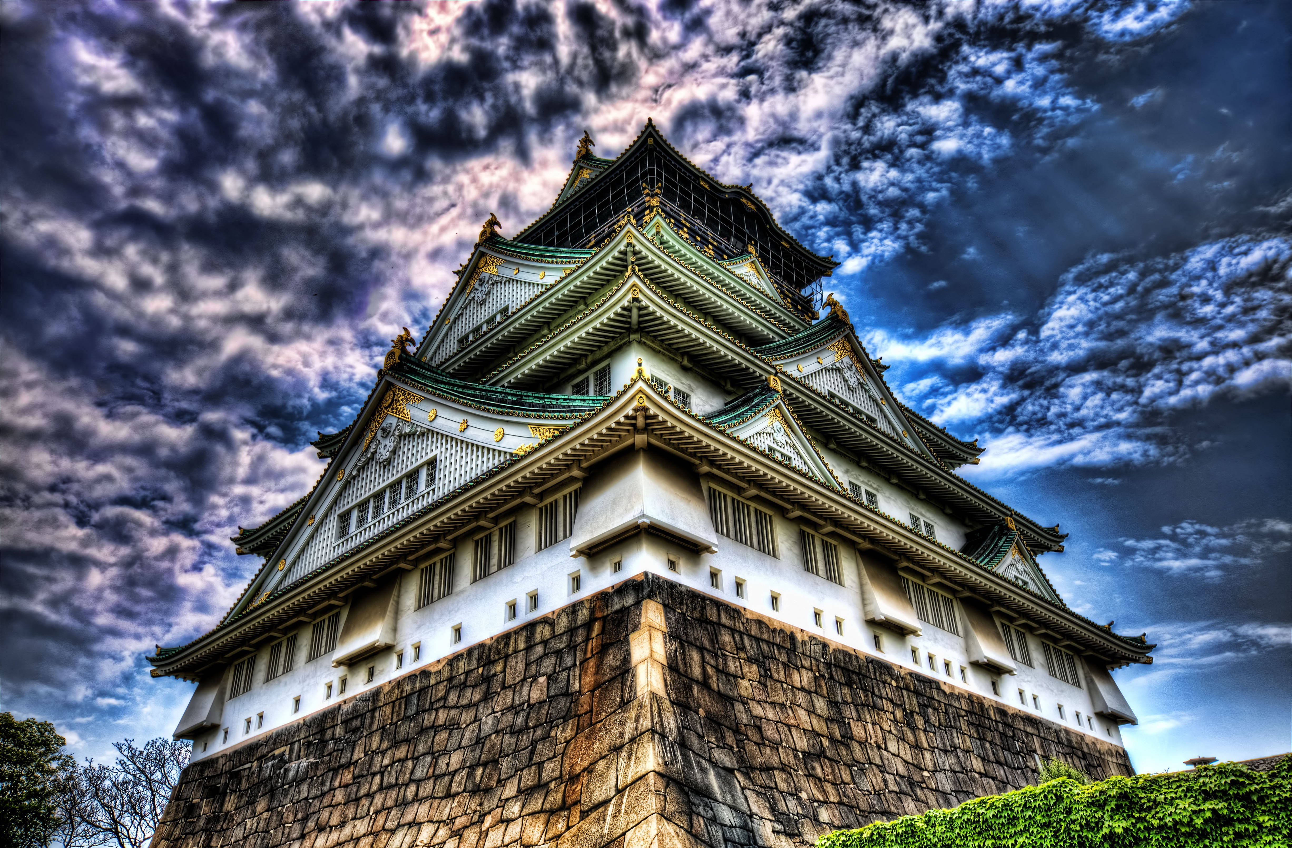 大阪城低角度拍攝 壁紙 — 一座雄偉的日本城堡塔樓，擁有綠色瓦片屋頂和白色牆壁，在戲劇性的多雲天空下巍然聳立。