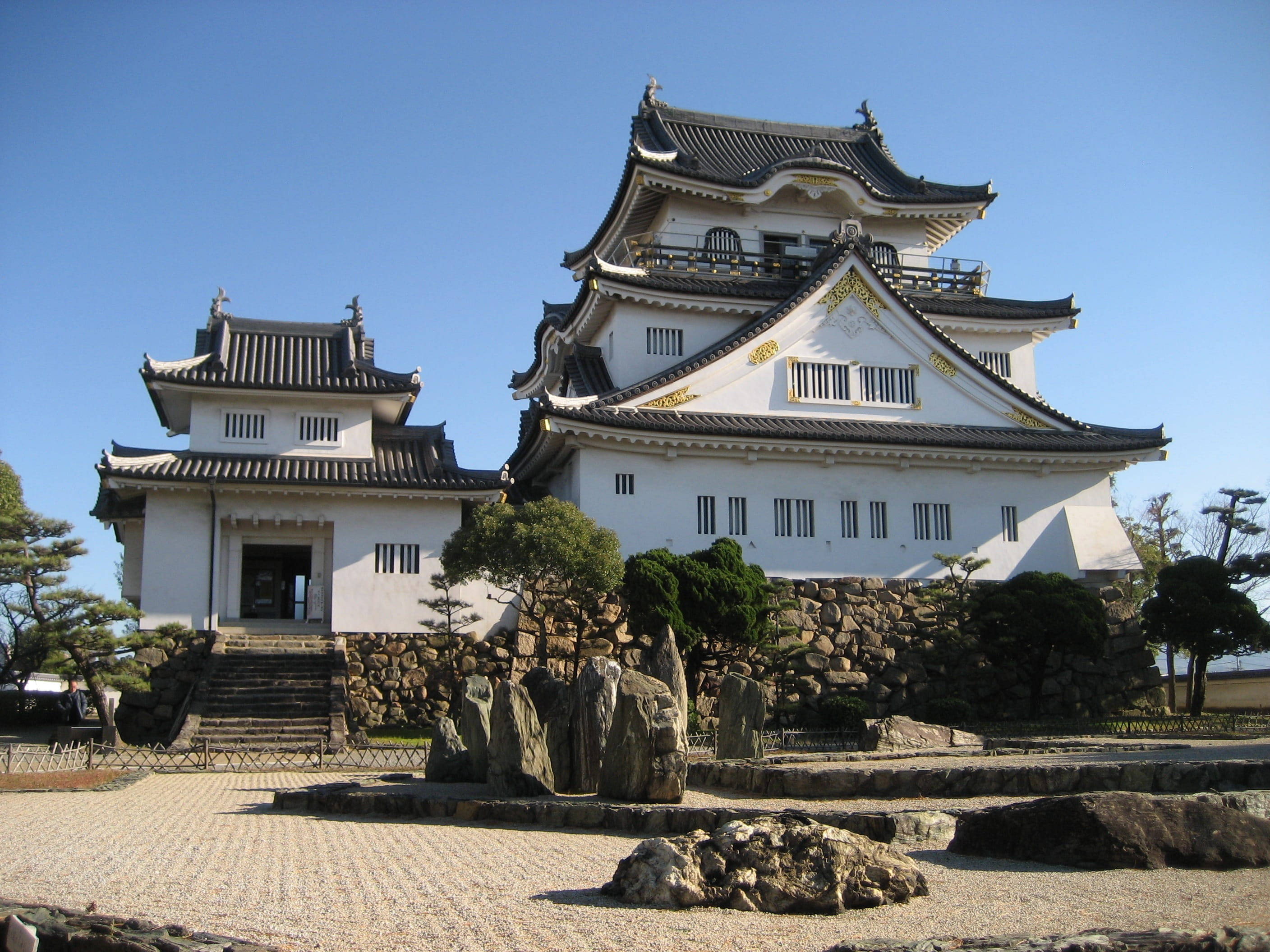 大阪城前的藝術岩石景觀 壁紙 — 一座宏偉的傳統日本城堡，白色牆壁和深色瓦片屋頂，在晴朗的藍天下，周圍是修剪整齊的花園和石徑。
