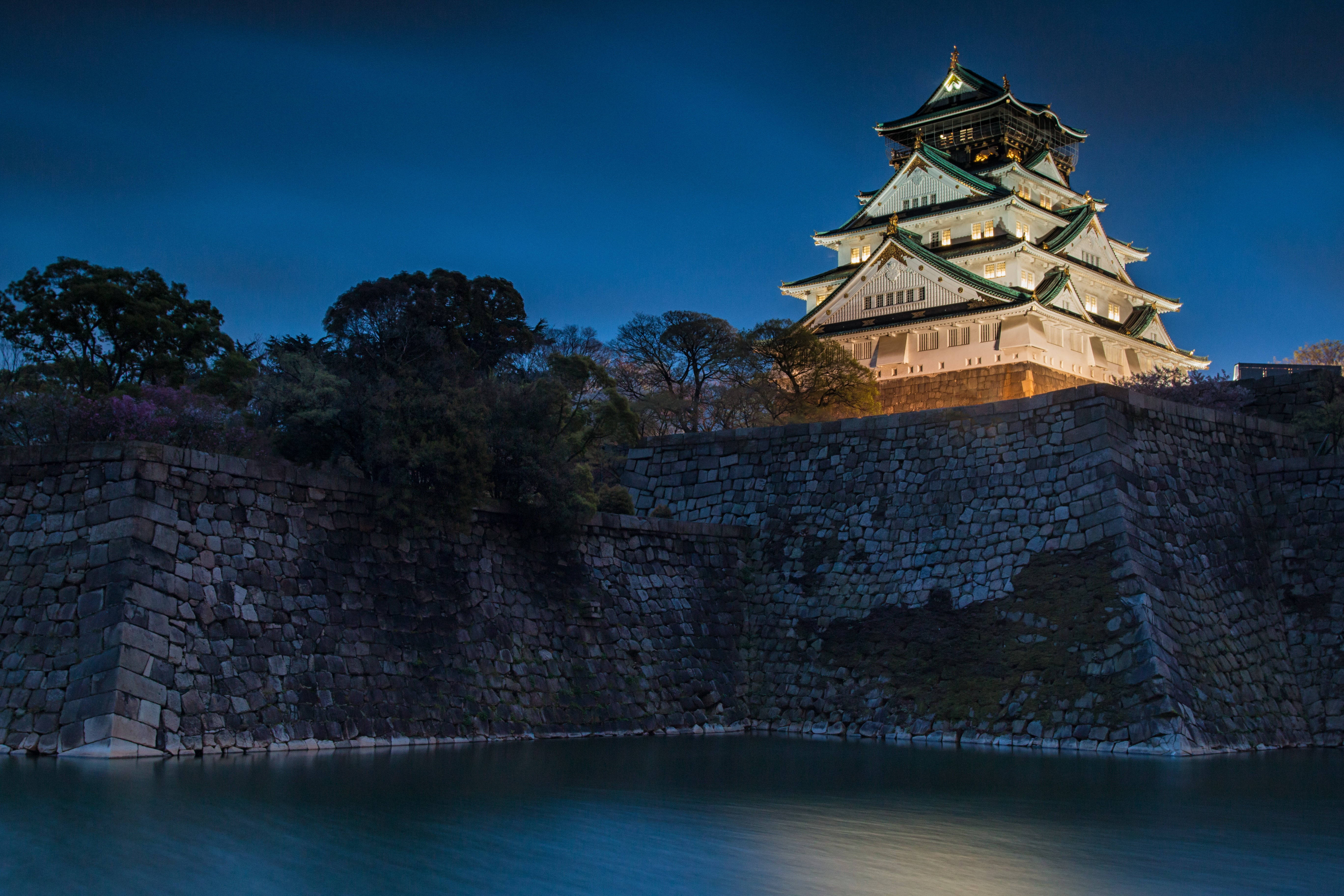 大阪城夜景 壁紙 — 一座雄偉的燈光璀璨的日本城堡在深藍色的暮色天空下矗立在平靜的護城河上。