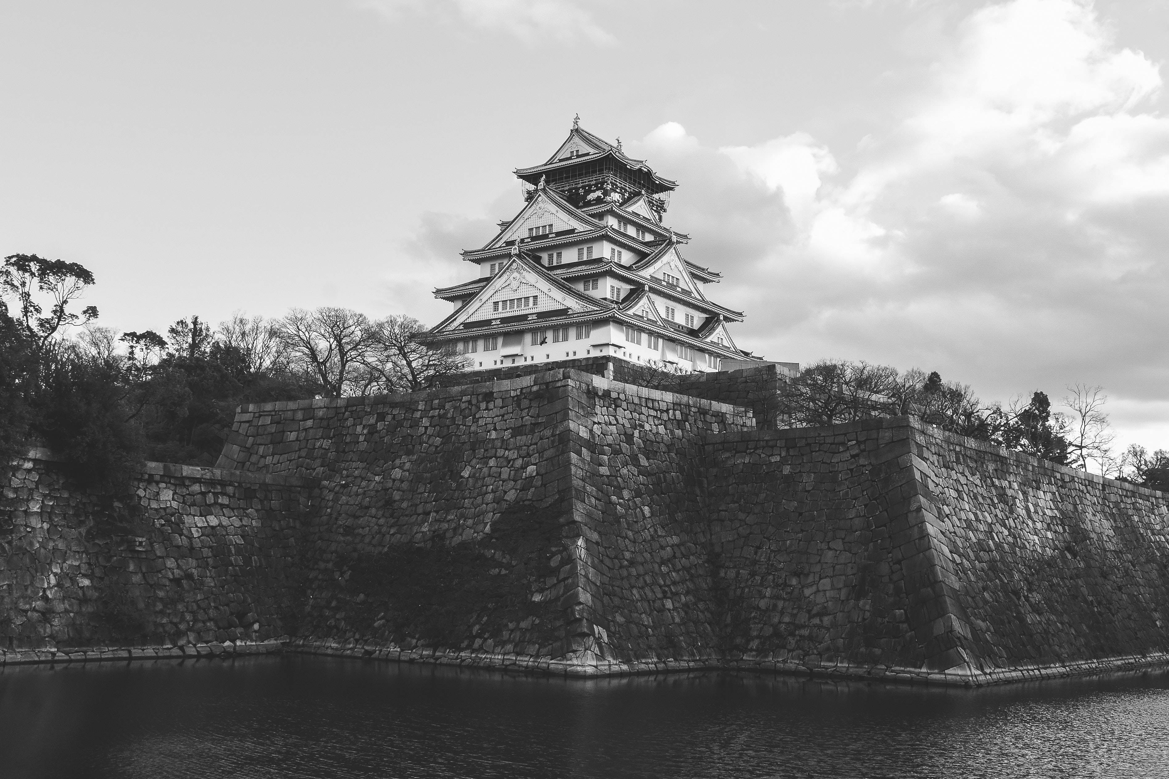 大阪城歷史氛圍 壁紙 — 一座雄偉的黑白城堡在陰天之下聳立於石牆和寧靜的護城河之上。