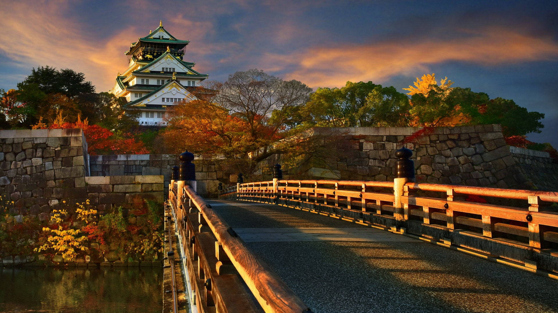 大阪城與極樂橋 壁紙 — 一條木橋通往日落時分雄偉的日本城堡，周圍有鮮豔的秋葉和溫暖的金色光芒。