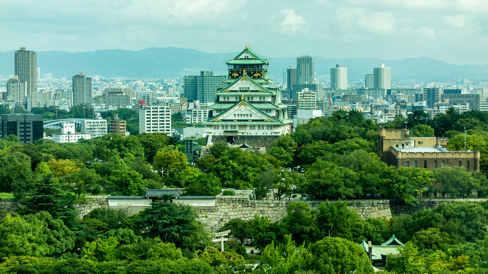 大阪城與綠意城市 壁紙 — 一座擁有綠色屋頂的宏偉傳統日本城堡坐落在鬱鬱蔥蔥的山丘上，俯瞰著多雲天空下的現代城市景觀。