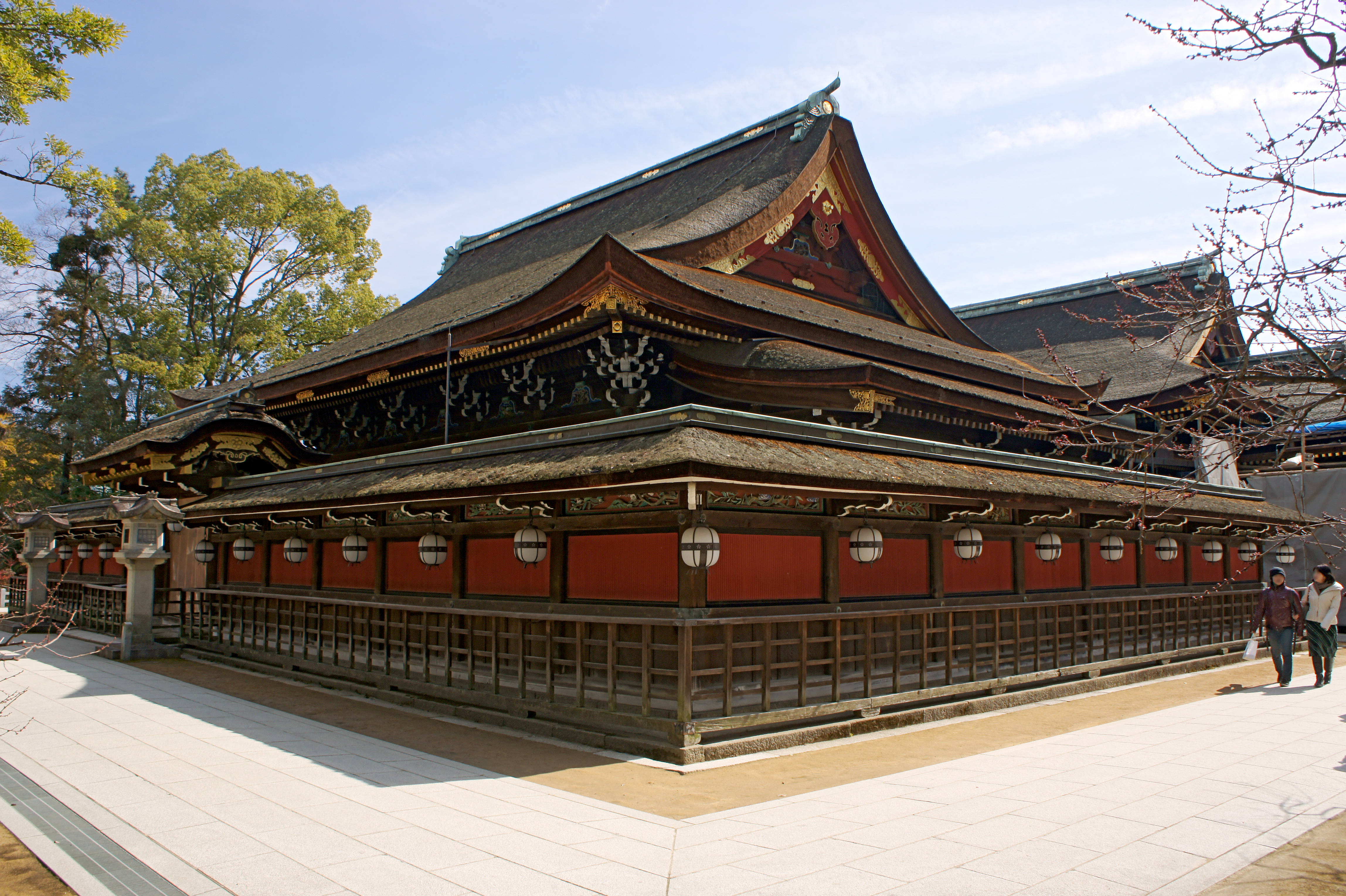 大阪天滿宮 壁紙 — 傳統日本神社，紅色牆壁和彎曲的深色屋頂，在晴朗的藍天下，有綠樹和兩個人在附近行走。