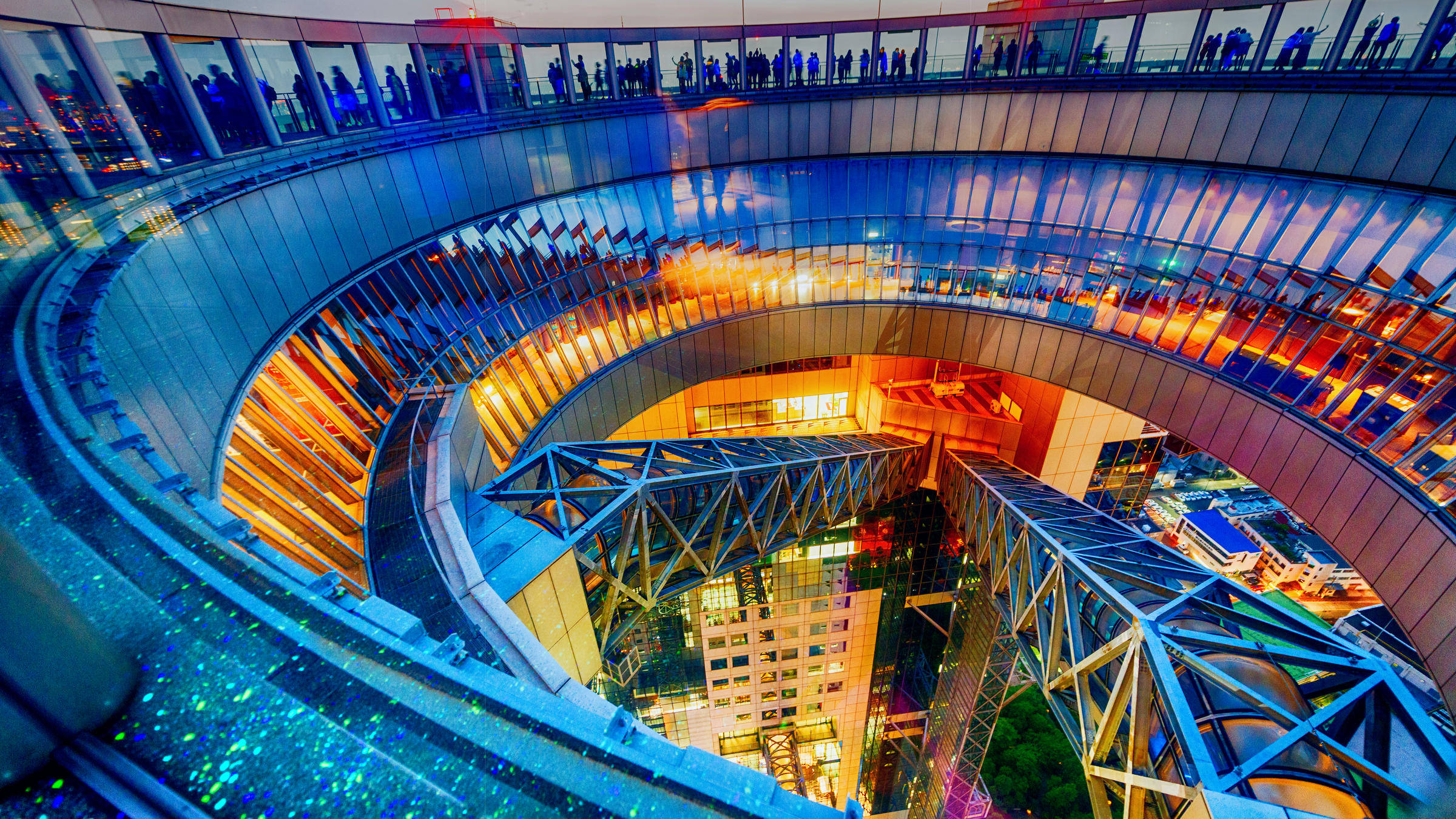 大阪天空大樓日落景色 壁紙 — 黃昏時分的未來主義螺旋玻璃步道，帶有發光的藍色和橙色燈光以及人物剪影。