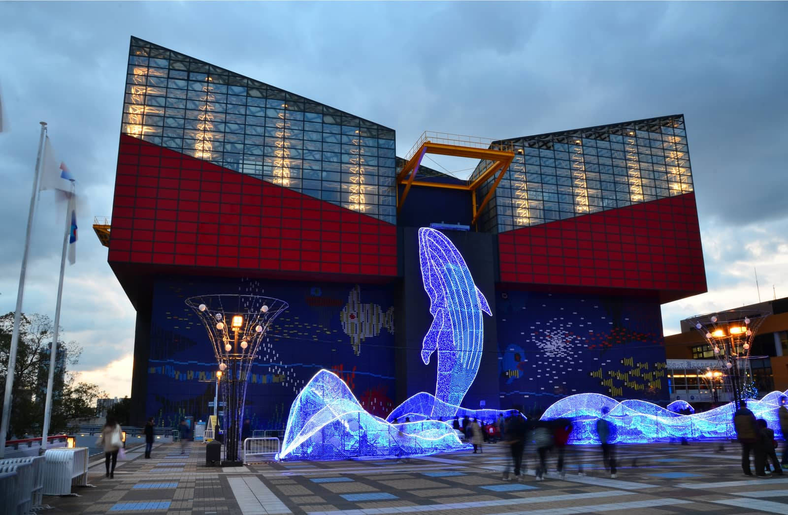 大阪海遊館水族館公園 壁紙 — 一座現代建築，紅色和玻璃外牆在黃昏時亮起，前面有一個發光的藍色鯨魚雕塑。