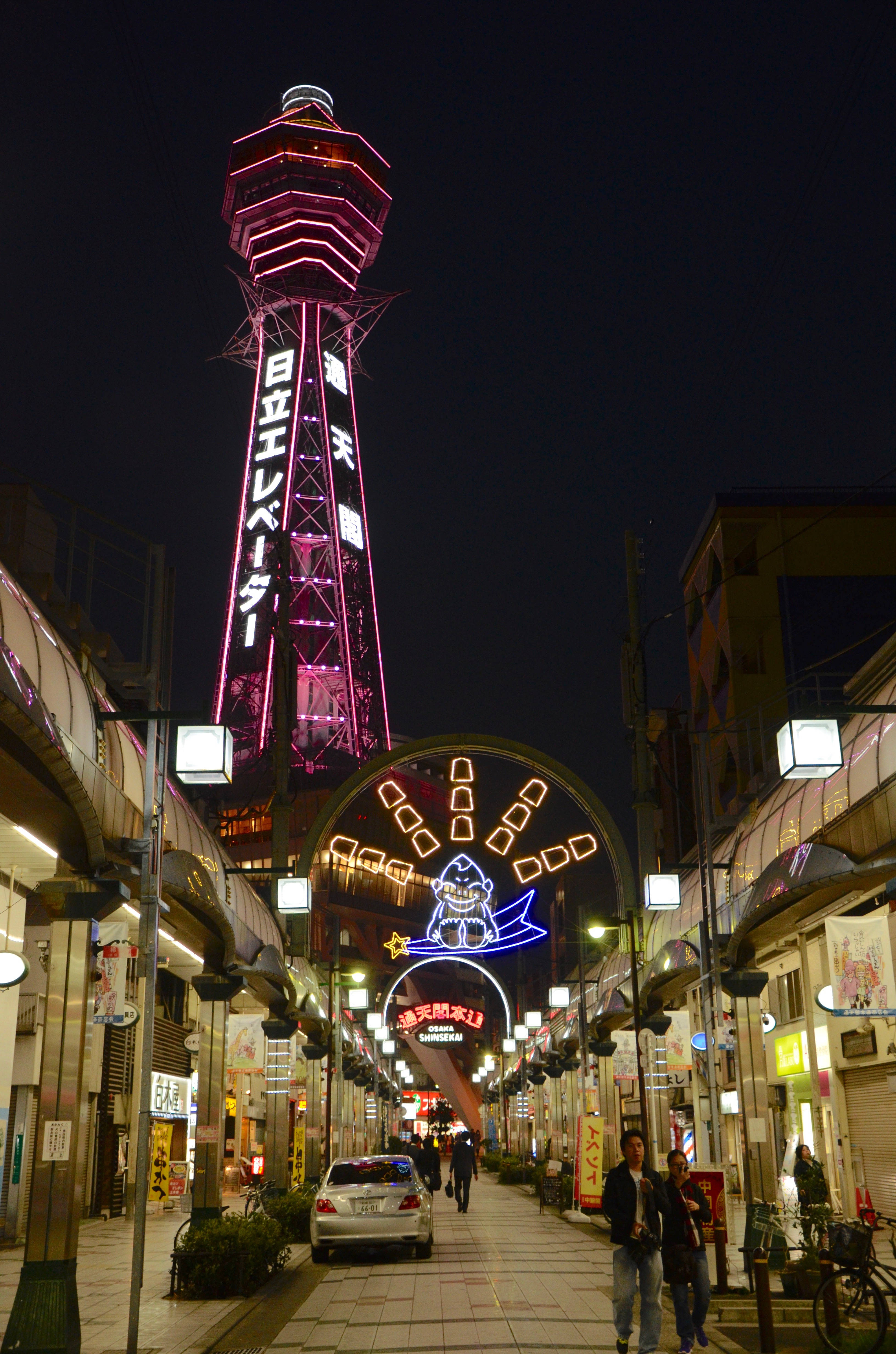 大阪通天閣 壁紙 — 日本城市街道的充滿活力的夜景，高聳的霓虹燈觀光塔和節日拱門在行人上方發光。