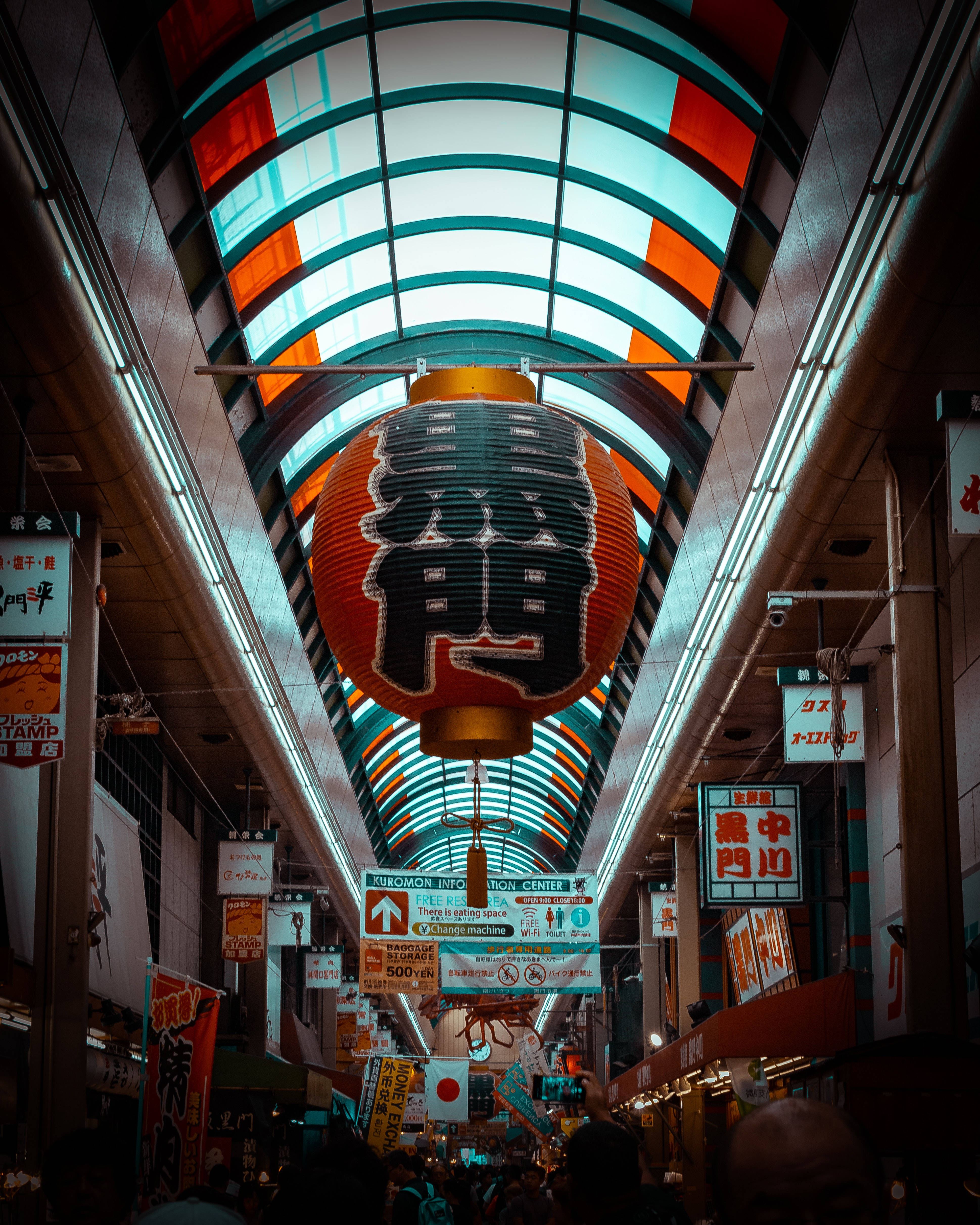 大阪黑門市場 壁紙 — 一個帶有黑色漢字的鮮紅色燈籠懸掛在熱鬧的有蓋拱廊中央，拱廊有色彩繽紛的拱形天窗和發光的招牌。