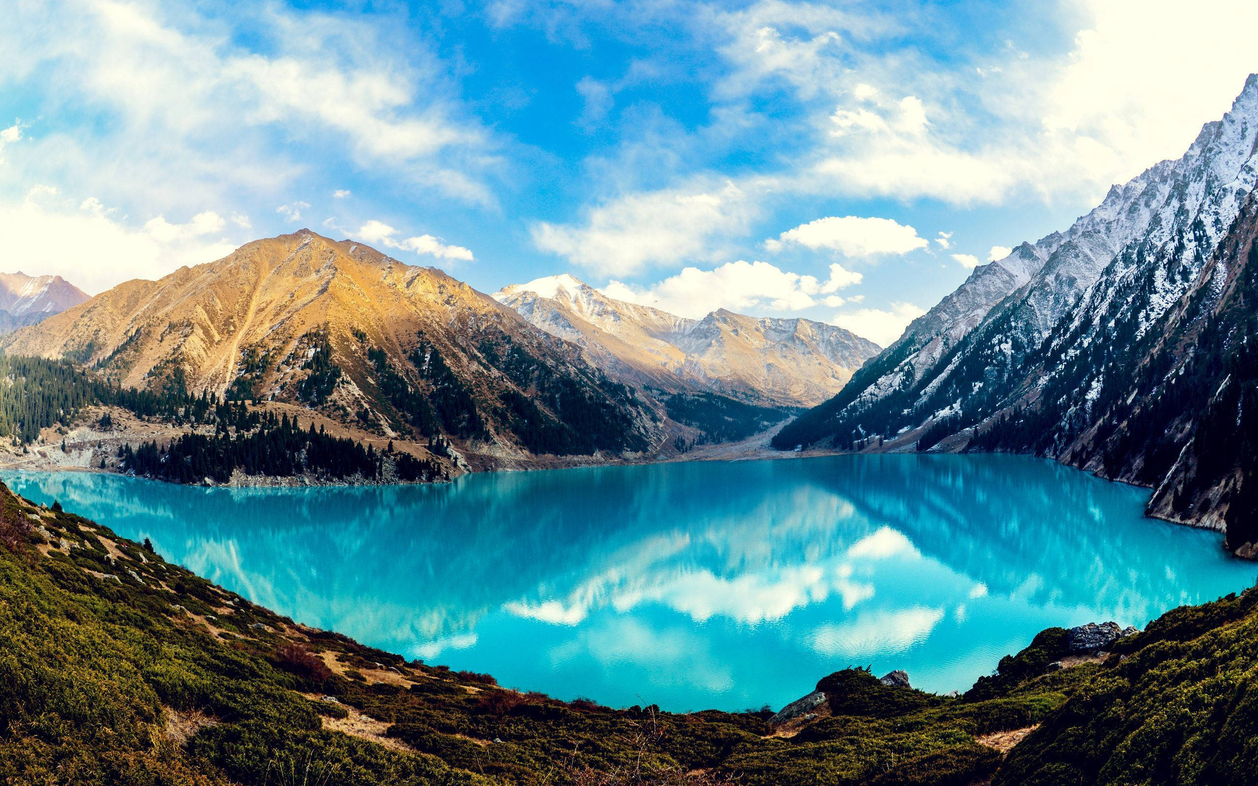 大阿拉木圖湖全景 壁紙 — 一個令人驚嘆的高山湖泊，擁有充滿活力的碧綠海水，在明亮的藍天和白雲下映照著白雪覆蓋的山峰。