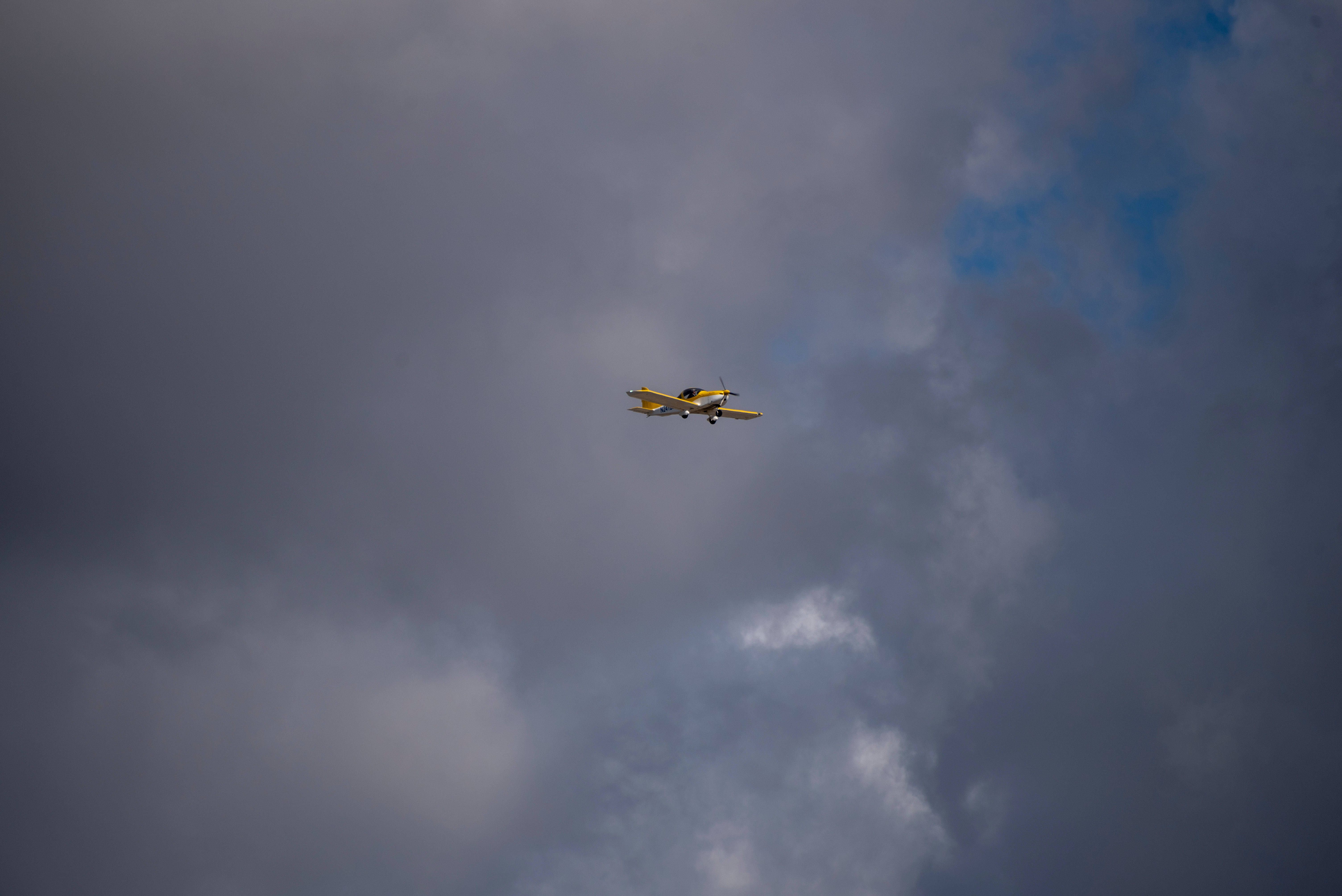 天空中的自由：黃色小型飛機高飛 壁紙 — 一架黃色小飛機在充滿深灰色雲朵和藍色斑塊的戲劇性天空中翱翔。